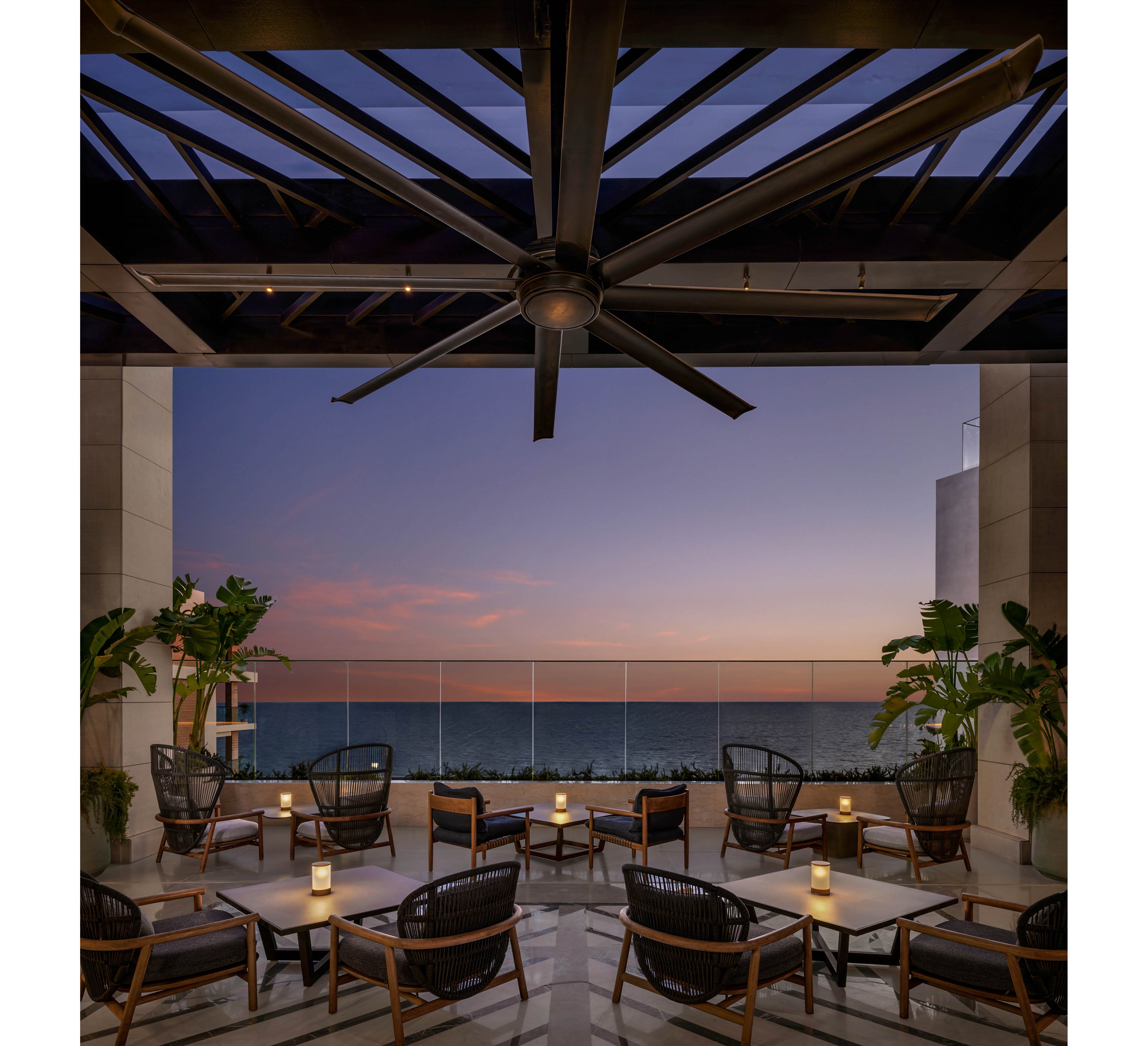 A contemporary open-air terrace lounge overlooks a calm ocean at dusk. Sleek armchairs and low tables are arranged in small seating groups beneath a large ceiling fan and exposed beams. Soft candle-like lights on each table create a warm, intimate glow against the cool twilight sky. Potted tropical plants frame the scene, adding greenery and a relaxed resort atmosphere.