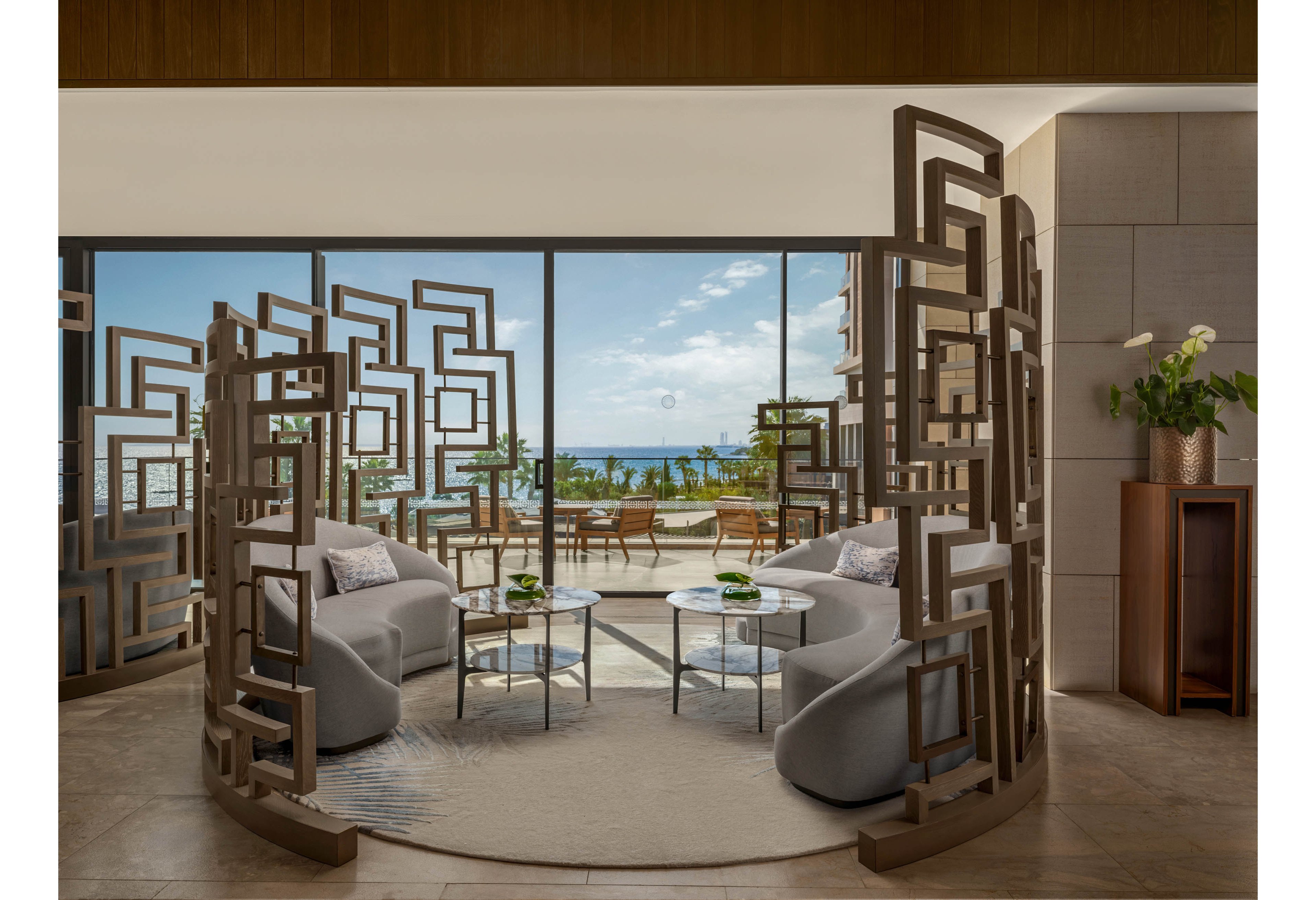 A contemporary lounge area features curved sofas arranged around glass coffee tables, framed by geometric wooden partitions. Floor-to-ceiling windows open onto a terrace with outdoor seating and lush greenery. Beyond the terrace, the ocean and blue sky create a serene coastal backdrop. Neutral tones, soft lighting, and clean lines give the space a luxurious, relaxing atmosphere.
