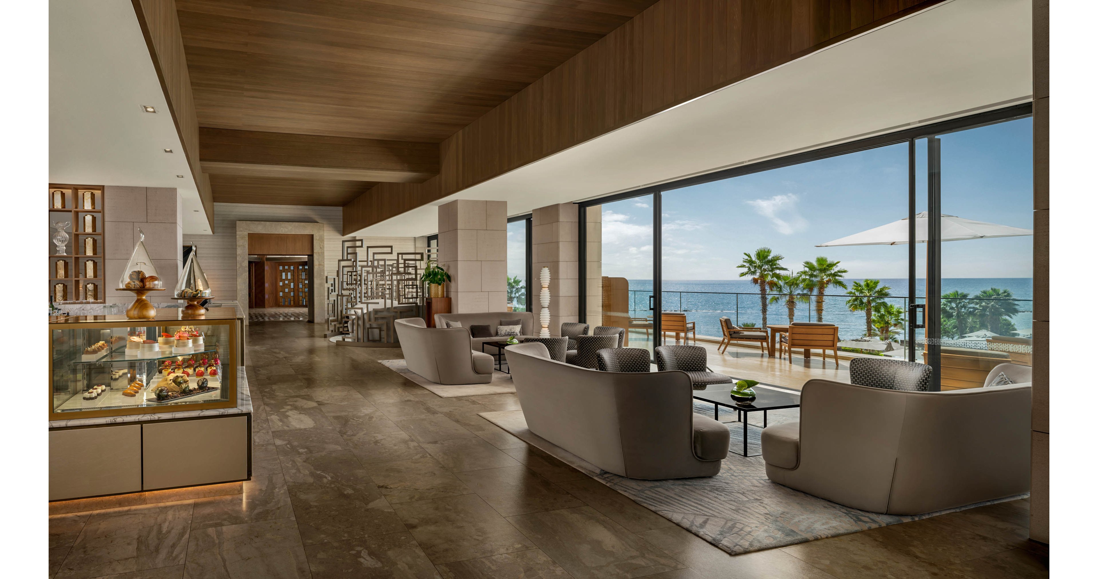 A contemporary hotel lounge features plush gray seating arranged around low tables facing large floor-to-ceiling windows. Beyond the glass, a sunny terrace with lounge chairs, umbrellas, palm trees, and a calm ocean view creates a resort-like atmosphere. Inside, warm wood ceilings, stone floors, and soft lighting contribute to a sophisticated yet relaxed ambiance. A glass display case with desserts or pastries adds a touch of hospitality near the seating area.