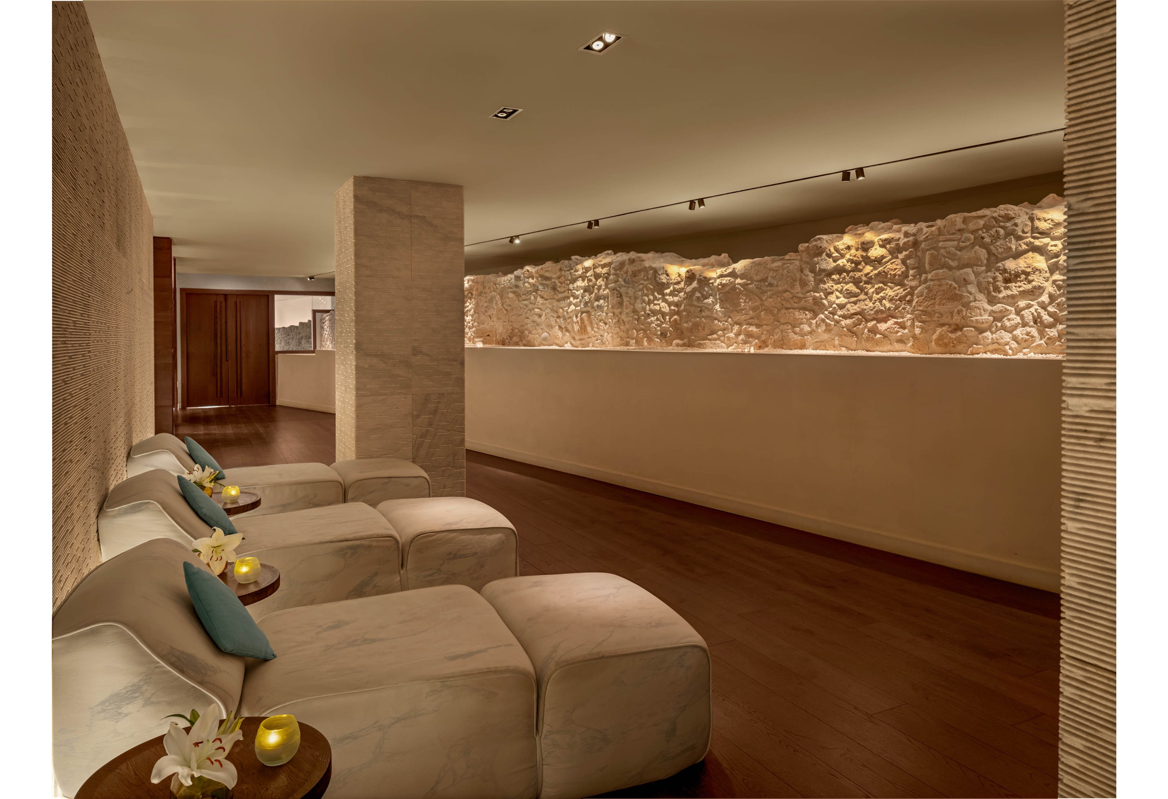 A serene spa lounge features a row of plush white loungers arranged along a warm wooden floor. Soft, indirect lighting highlights a textured stone wall, creating a calm and intimate atmosphere. Small side tables hold candles, flowers, and decorative items, adding subtle accents of color. The overall setting feels quiet and inviting, ideal for relaxation and wellness.