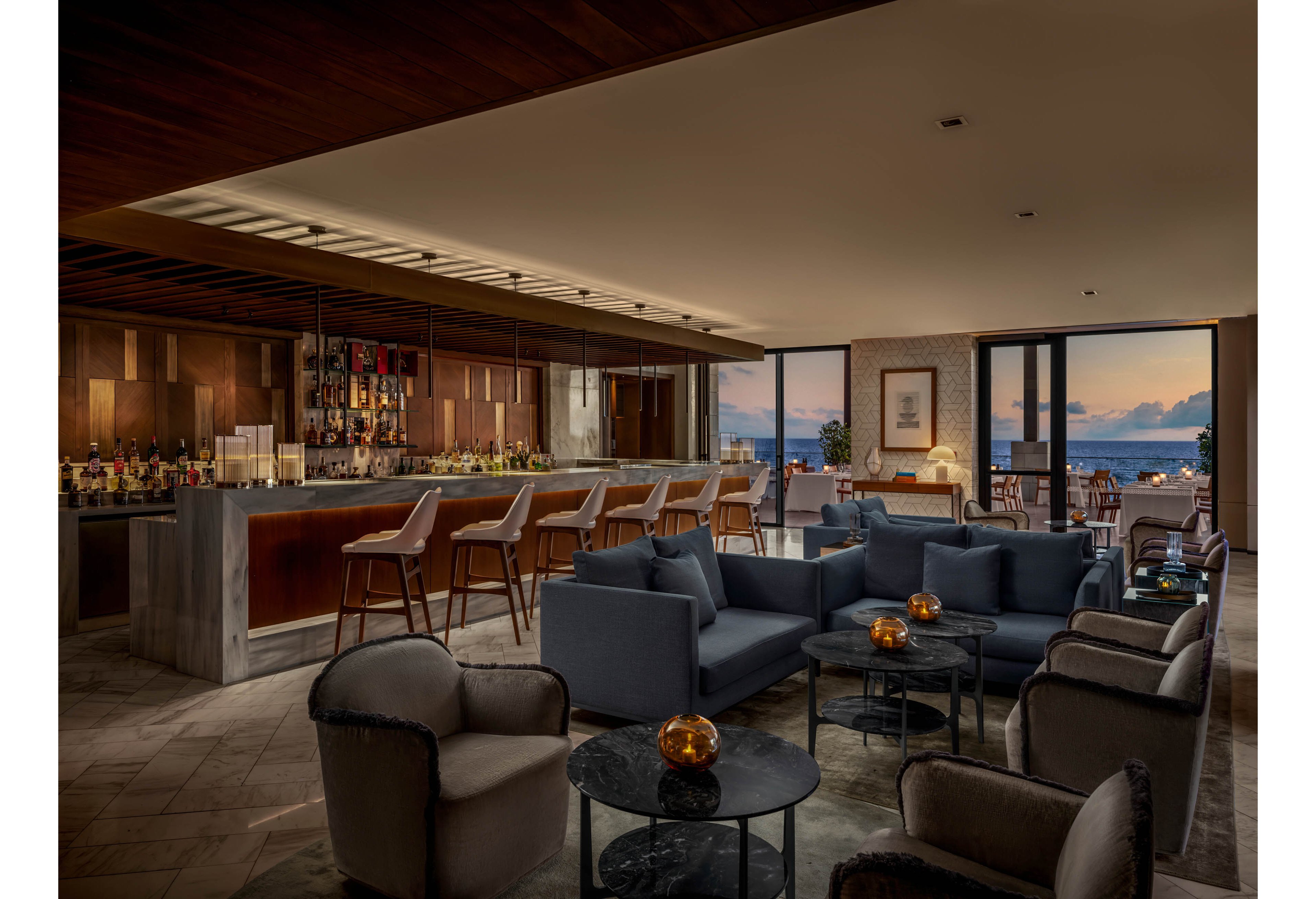 A contemporary hotel lounge bar features plush armchairs and sofas arranged around small marble tables. A sleek bar with high stools and a well-stocked back bar runs along one side of the room. Warm ambient lighting and candlelit tables create an inviting, intimate atmosphere. Large windows open to a terrace with views of the ocean and a colorful sunset sky.