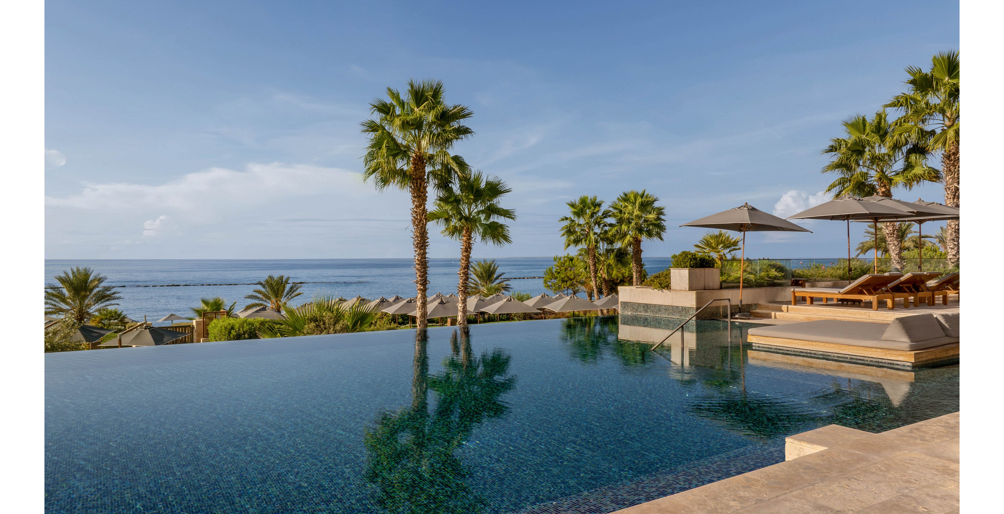 A serene infinity pool stretches toward a calm ocean horizon, creating a seamless blend between water and sky. Palm trees line the edge of the resort, adding a lush tropical feel to the scene. Sun loungers and umbrellas sit neatly arranged on a stone terrace beside the pool, suggesting a luxurious vacation setting. The clear blue water and soft natural light evoke a peaceful, relaxing atmosphere.