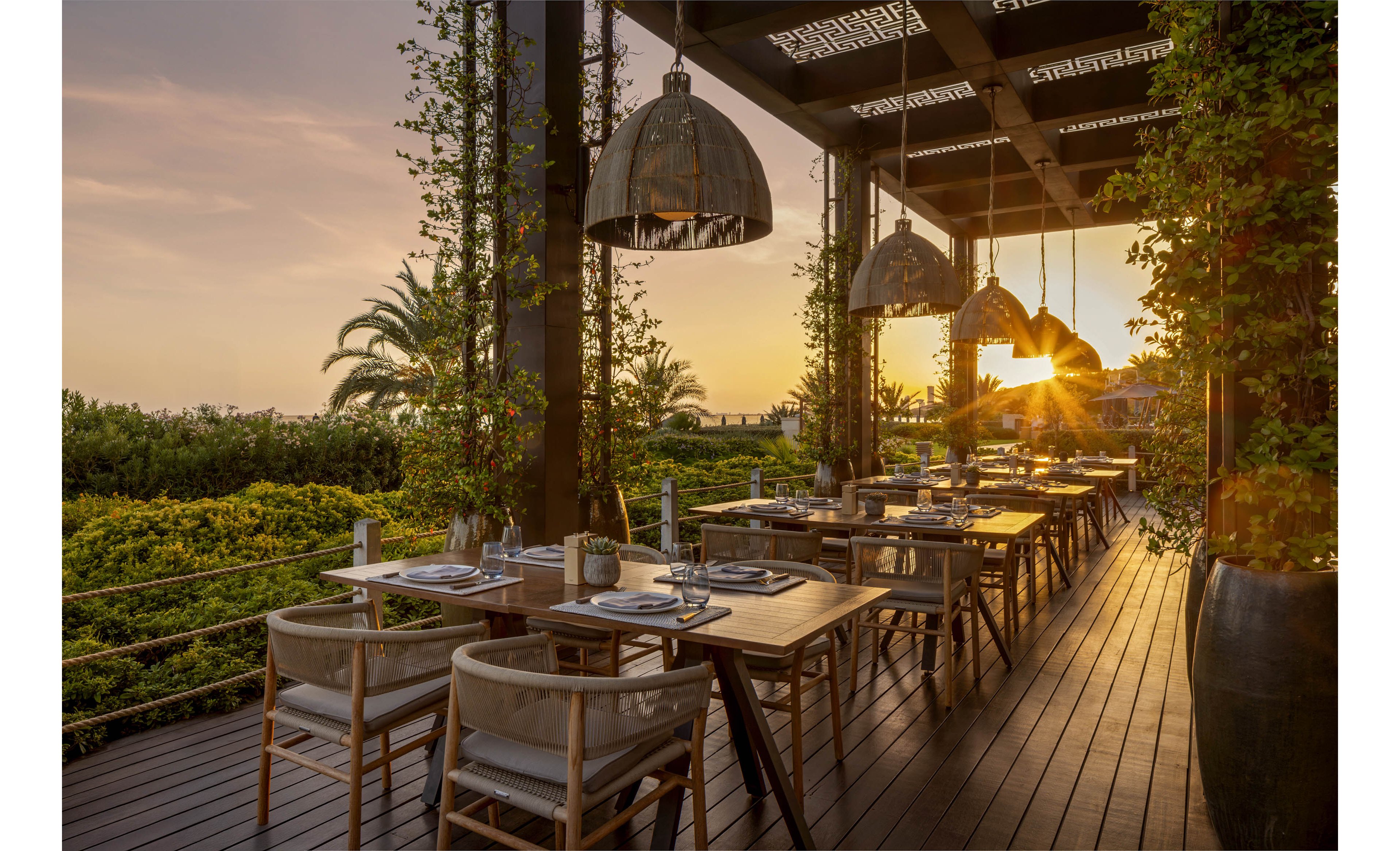 An elegant open-air restaurant terrace is set for dinner at sunset. Wooden tables and cushioned chairs line the deck beneath a modern pergola with large hanging lamps. The warm sunlight filters through the structure, casting long shadows across the floor. Lush green foliage and a distant palm tree frame the view, creating a tranquil, upscale outdoor dining atmosphere.