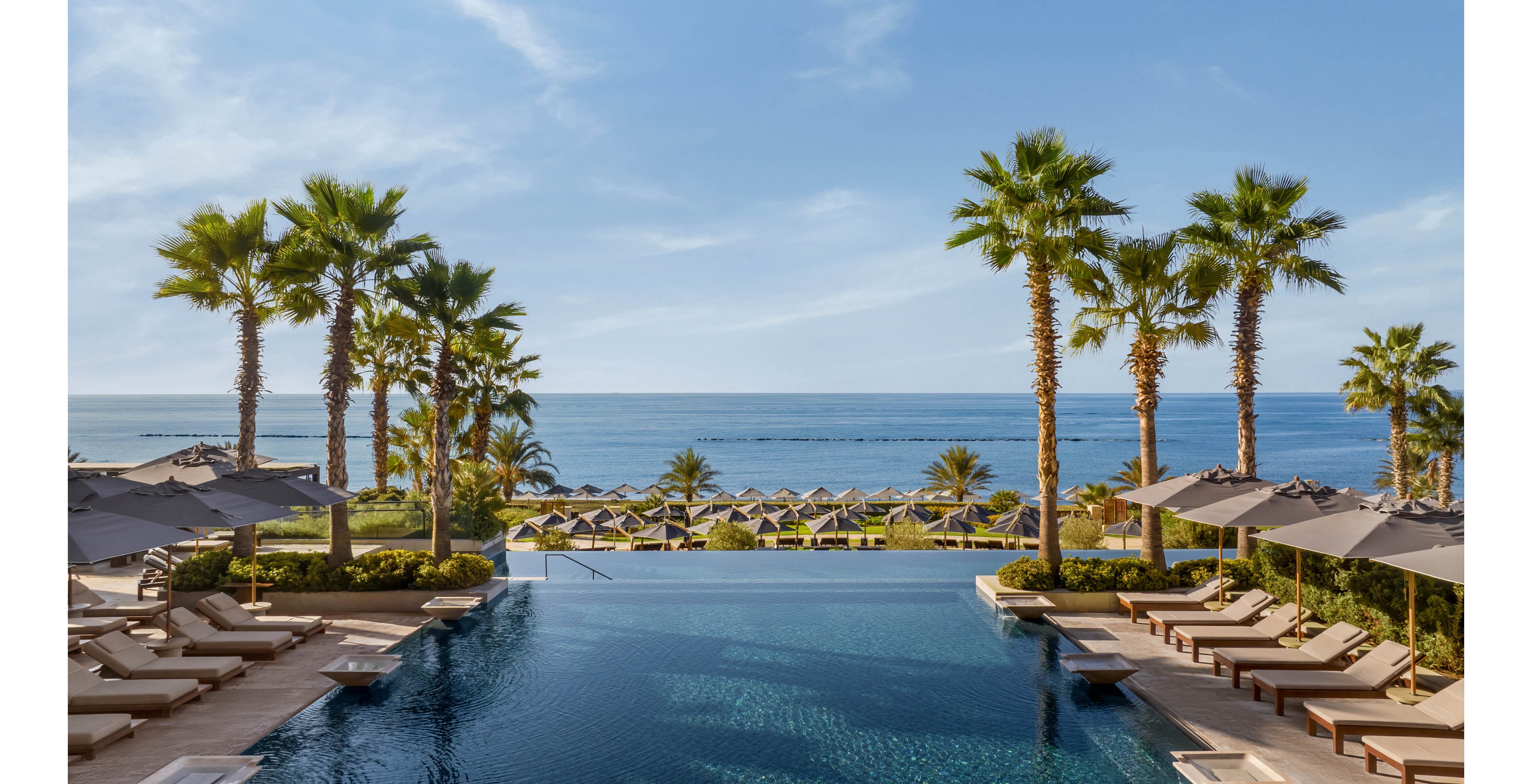 A serene infinity pool stretches toward a calm ocean horizon, framed by tall palm trees. Sun loungers and umbrellas line both sides of the pool, suggesting a luxurious resort setting. In the distance, rows of beach chairs and parasols sit on a manicured lawn leading to the shoreline. Clear blue skies and gentle waves create a tranquil, vacation-like atmosphere.