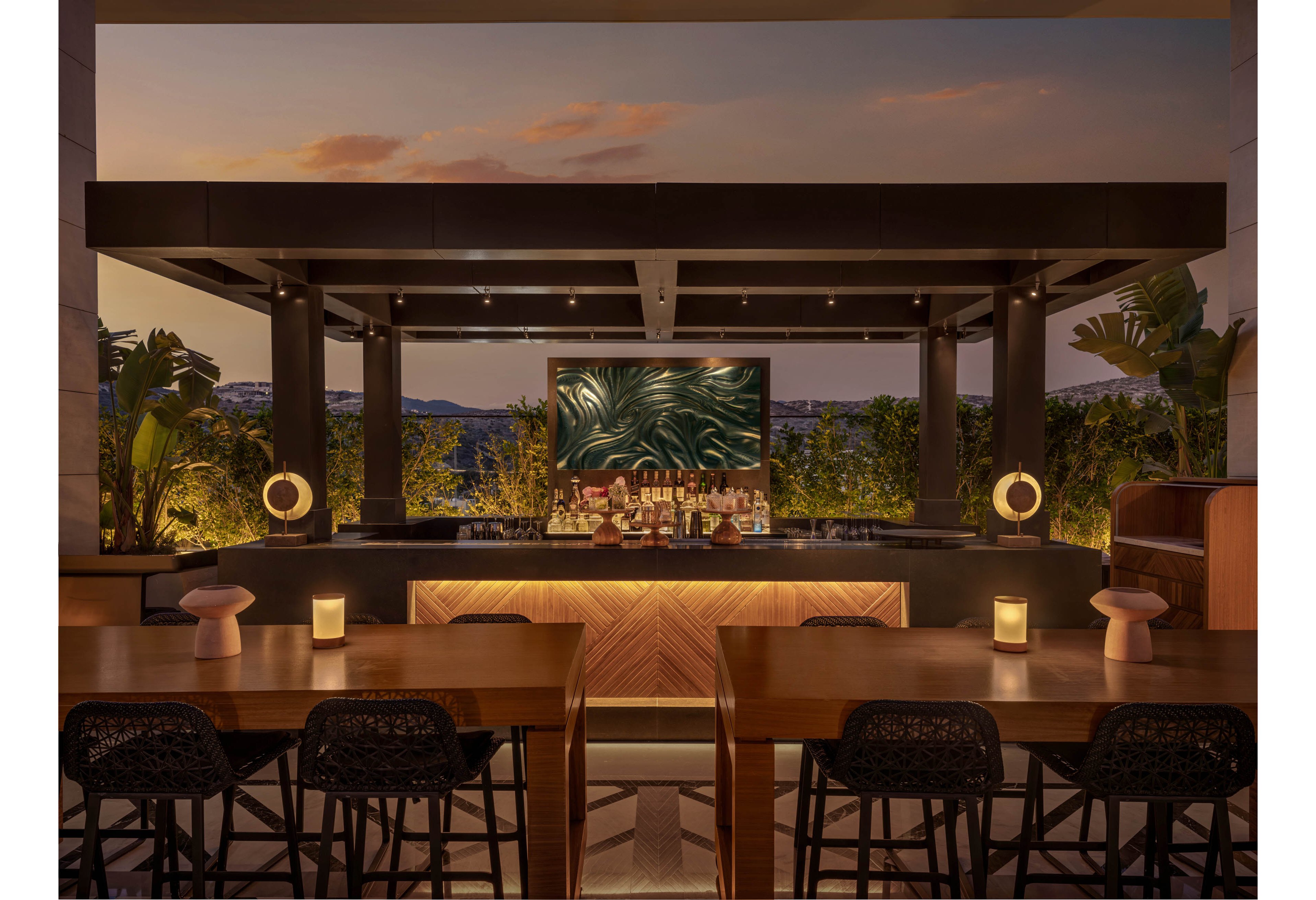 An elegant outdoor bar is set on a rooftop terrace at sunset, framed by a dark pergola structure. A long wooden bar counter with backlit panels faces several high stools and communal tables in the foreground. Warm ambient lighting from table candles and circular lamps creates a cozy, intimate atmosphere. Lush greenery and distant hills form a serene backdrop behind the bar, while a large abstract artwork adds visual interest.