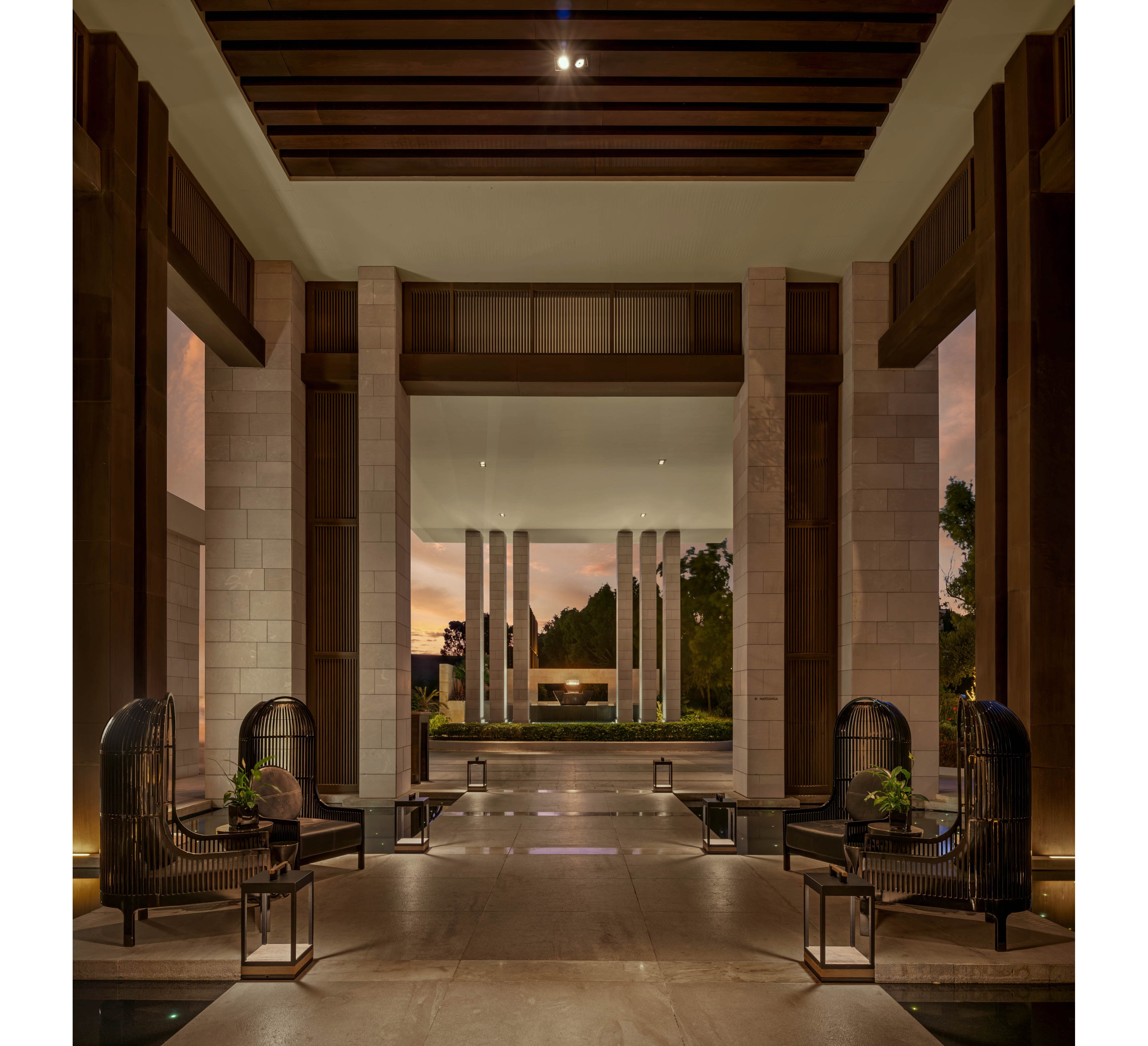 A grand open-air lobby features tall columns and a high ceiling with dark wooden beams. Elegant wicker lounge chairs with cushions and small side tables line the polished stone floor. Lanterns placed along the walkway create a warm, inviting glow as evening light filters in from outside. In the distance, a modern water feature and trees frame the tranquil outdoor view.