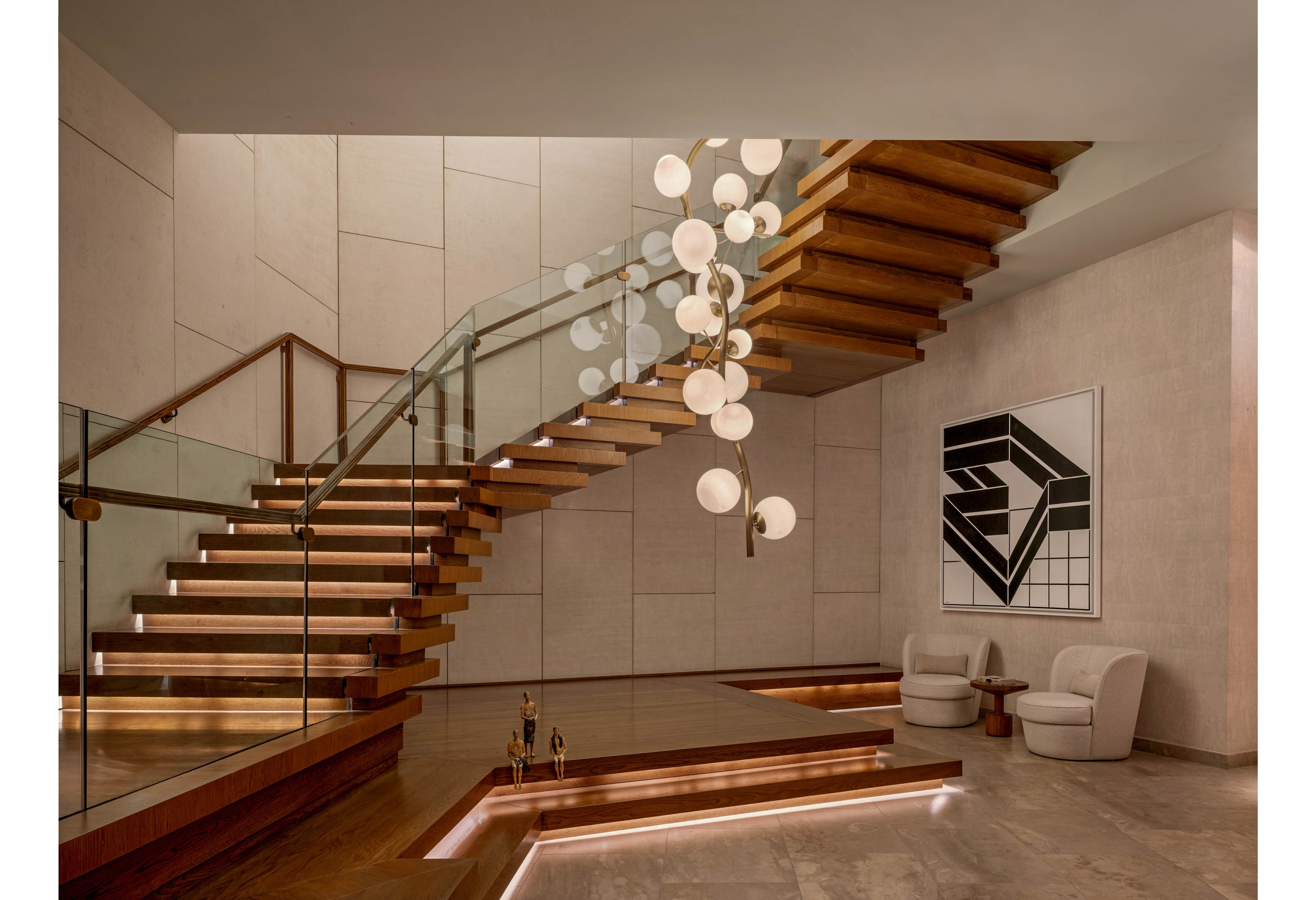A contemporary interior features a dramatic floating wooden staircase with glass railings and integrated step lighting. A sculptural chandelier with spherical lights hangs in the center of the stairwell, creating a focal point. The space includes a recessed seating area with two modern armchairs and a small side table. Large tiled walls and a geometric black-and-white artwork contribute to the sophisticated, minimalist aesthetic.