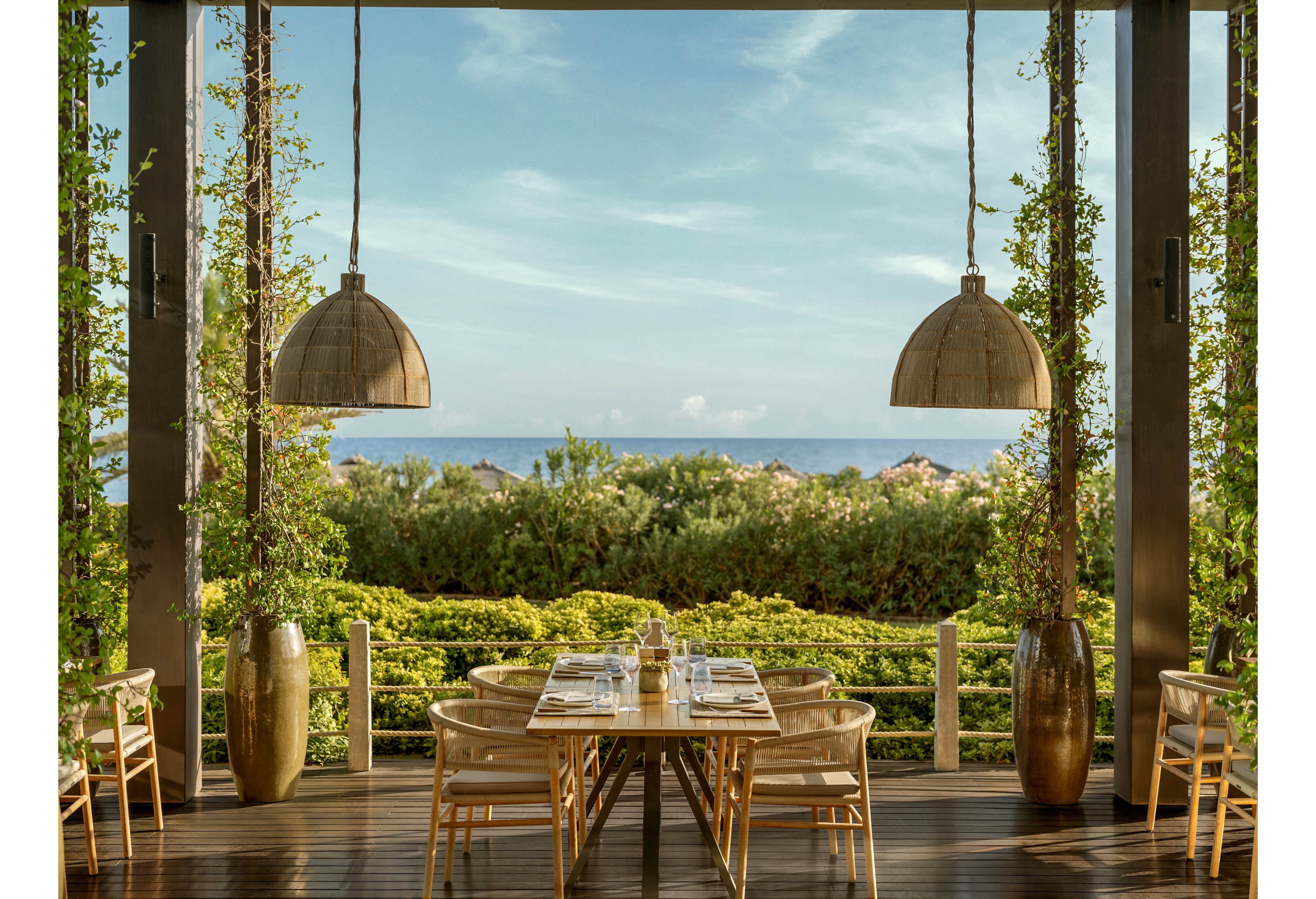 An elegant open-air restaurant terrace features a wooden table set for dining with glassware and plates. Light wooden chairs surround the table, positioned under two large hanging dome lamps. Lush green foliage and potted plants frame the view, leading the eye toward the distant ocean and blue sky. The scene feels tranquil and inviting, ideal for a relaxed coastal meal.