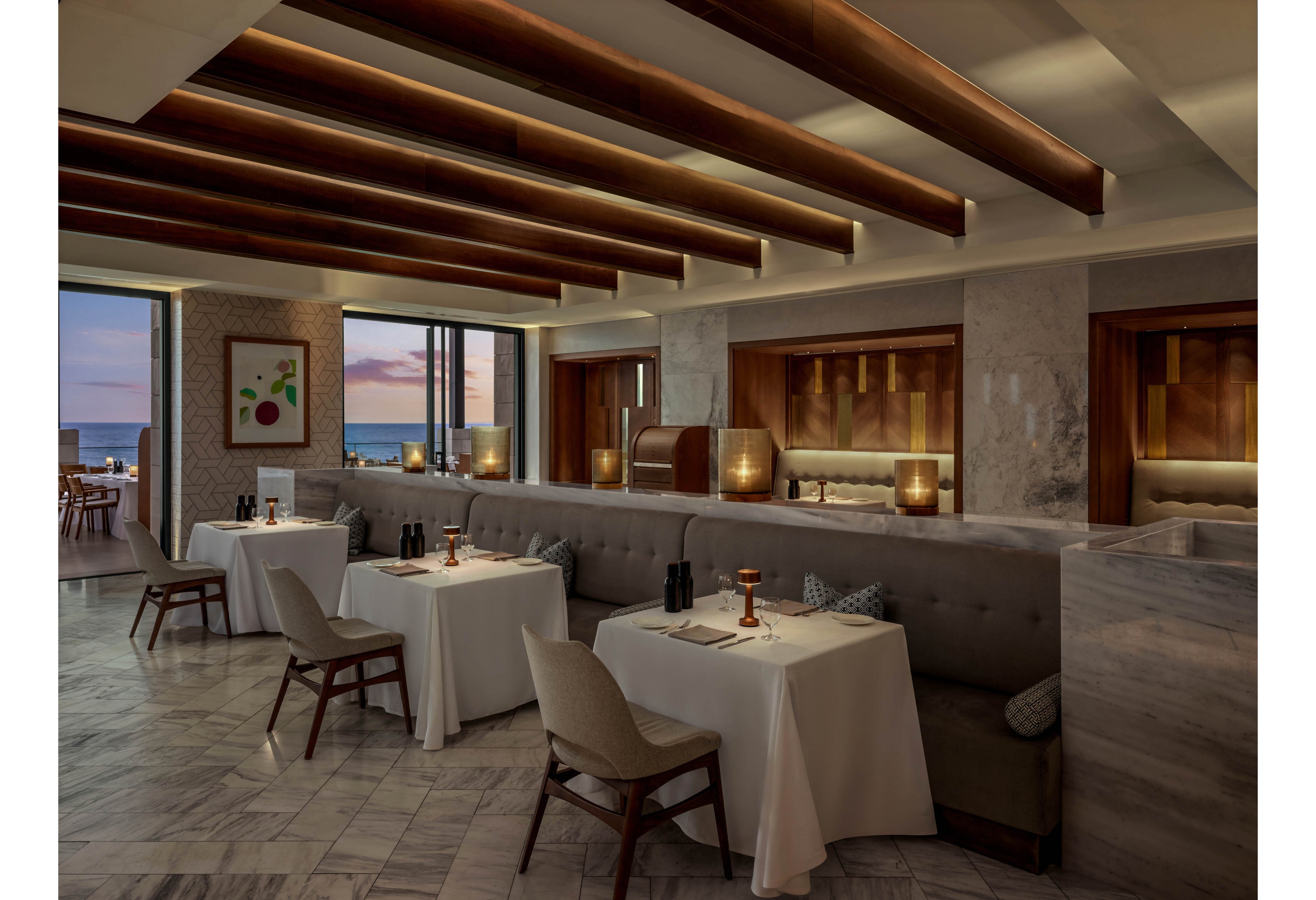 A refined restaurant dining room features neatly arranged tables with white linens and modern chairs. Soft, warm lighting from table lamps and wall fixtures creates an intimate atmosphere. Large windows reveal an ocean view and colorful sunset sky in the background. Contemporary artwork and wood accents complement the marble floors and neutral-toned seating, emphasizing a luxurious coastal setting.