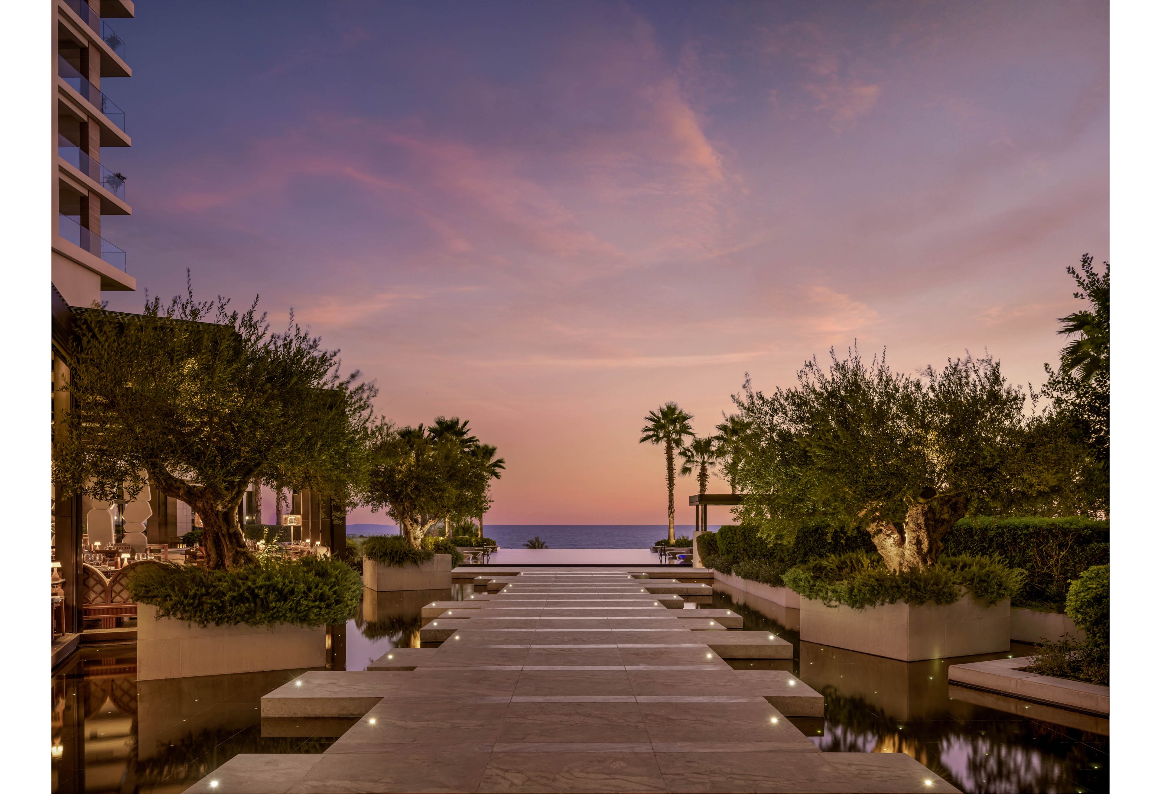 A contemporary resort walkway leads toward the sea under a colorful sunset sky. Wide stone steps are bordered by shallow reflecting pools and neatly trimmed trees in large planters. Palm trees frame the view, with a glimpse of the ocean horizon in the distance. Soft embedded lights along the path create a calm, luxurious atmosphere.