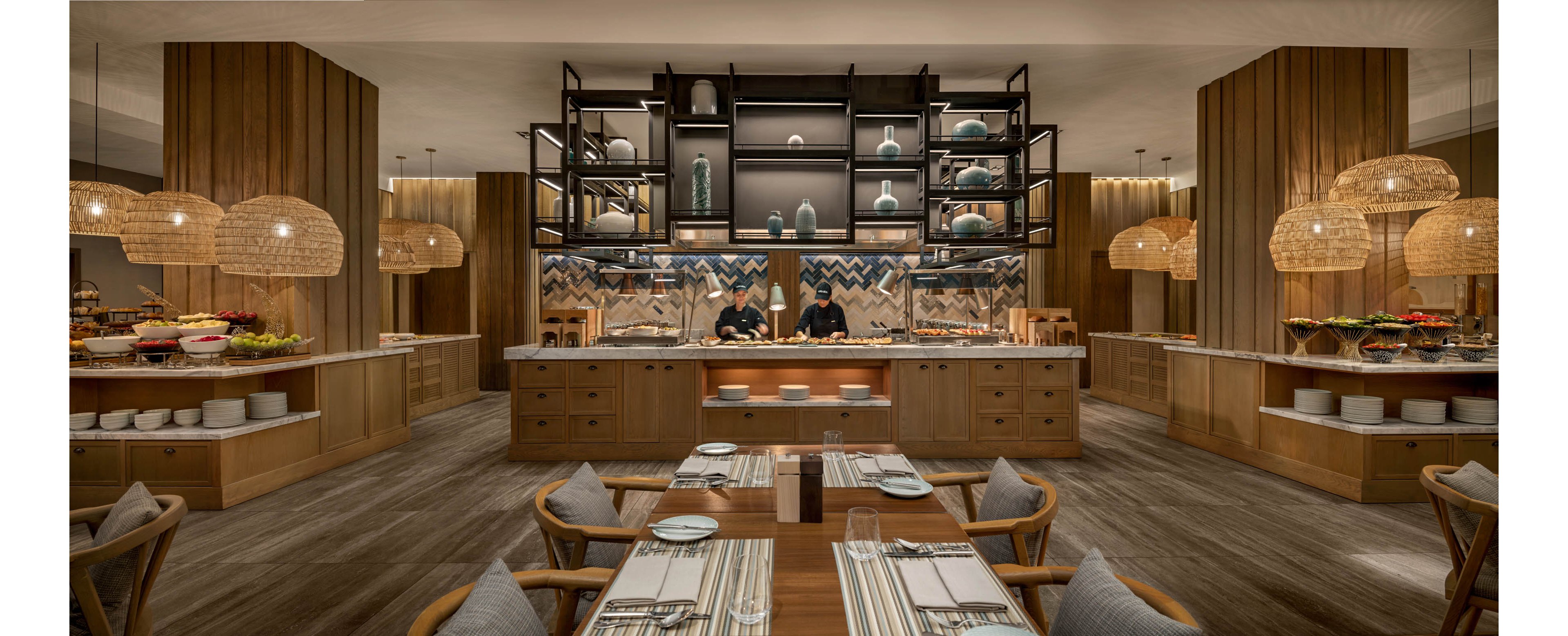 A contemporary restaurant interior features a central buffet counter with two chefs preparing food. Wooden tables and chairs are neatly arranged in the foreground, set with placemats, cutlery, and napkins. Warm pendant lights and wooden paneling create a cozy, upscale ambiance. Decorative shelving, ceramics, and patterned wall tiles add visual interest behind the serving area.