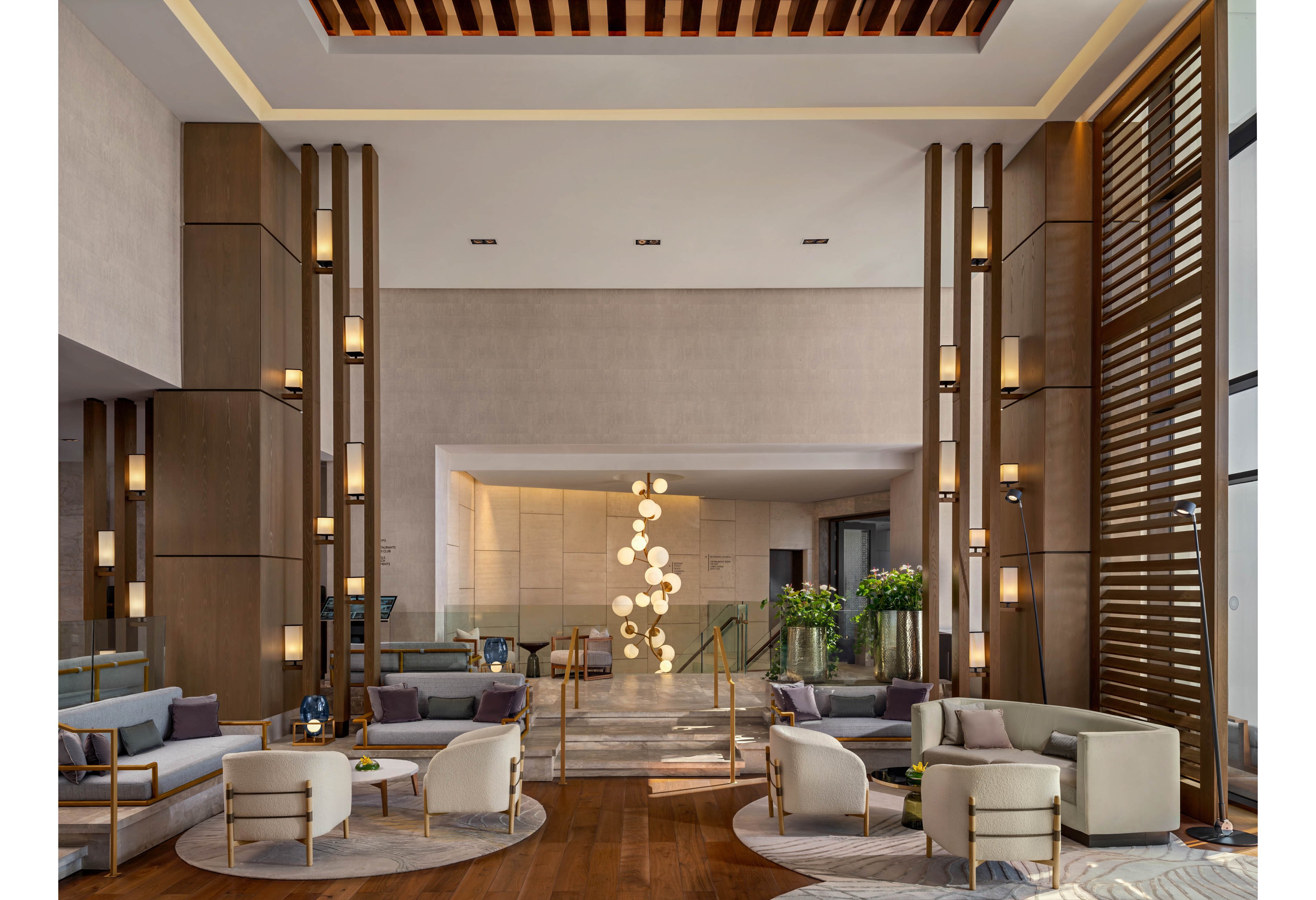 A spacious, contemporary hotel lobby features low lounge seating arranged around circular rugs. Tall wooden columns with integrated warm lights frame the space and draw the eye upward. A sculptural chandelier of glowing spheres hangs in the background near a staircase with glass railings. Neutral tones of beige, gray, and wood create a calm, sophisticated atmosphere accented by green plants.