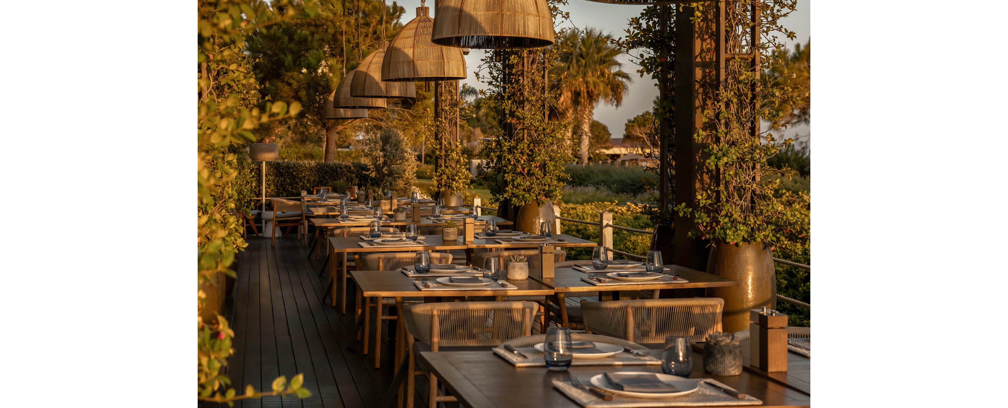 An elegant outdoor restaurant terrace is set for dining in the warm light of sunset. Wooden tables are neatly arranged in a row, each prepared with plates, cutlery, and glassware. Large woven pendant lamps hang above the tables, casting a soft glow over the scene. Lush greenery and trees surround the deck, creating a tranquil and inviting atmosphere.