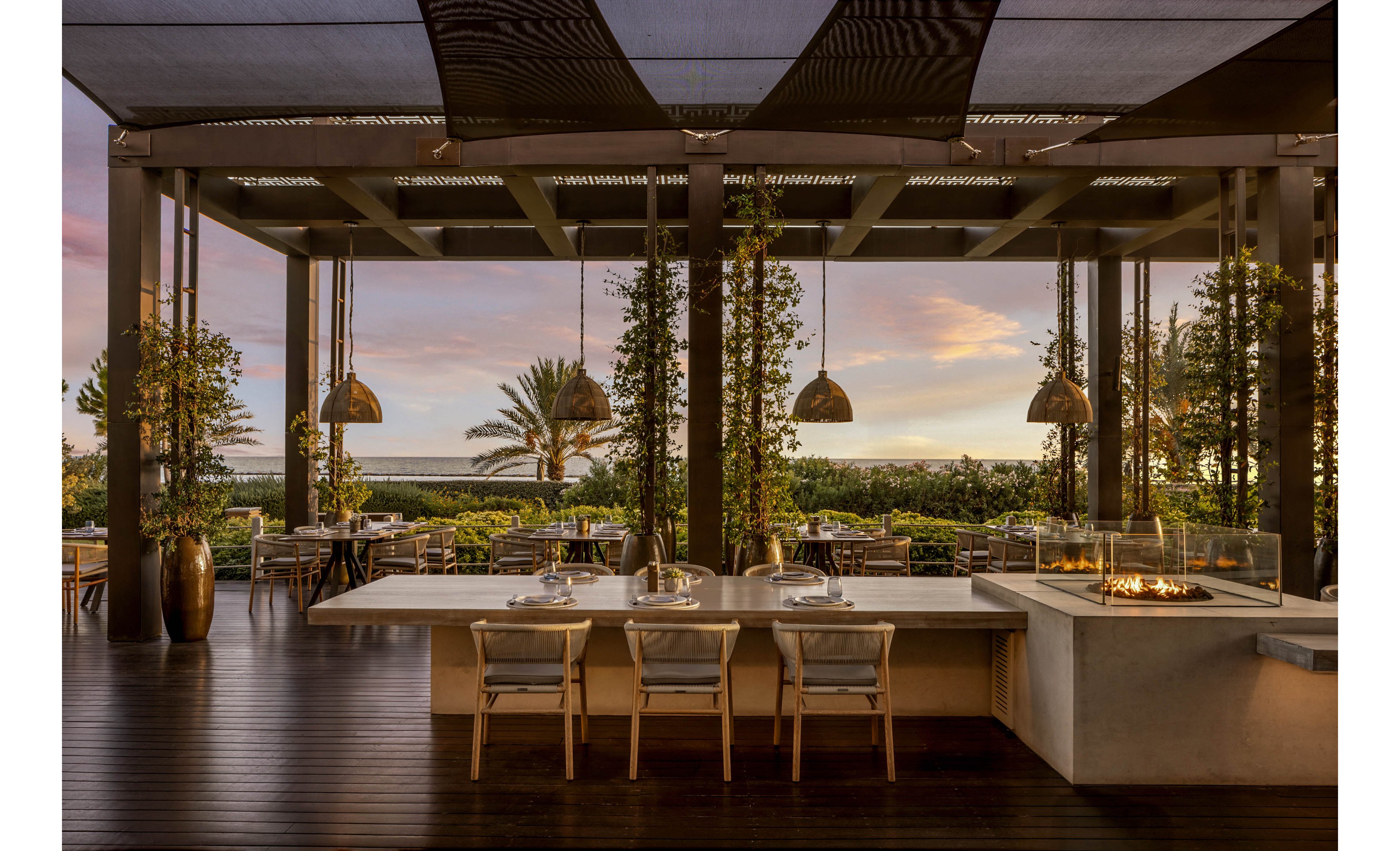 An elegant open-air restaurant terrace features a long communal table and modern chairs facing a sleek outdoor grill and fire feature. The space is framed by tall columns, hanging pendant lights, and vertical greenery that softens the contemporary architecture. Beyond the terrace, lush landscaping leads the eye toward palm trees and a distant ocean horizon under a pastel sunset sky. The warm lighting and natural materials create a relaxed yet sophisticated dining atmosphere.