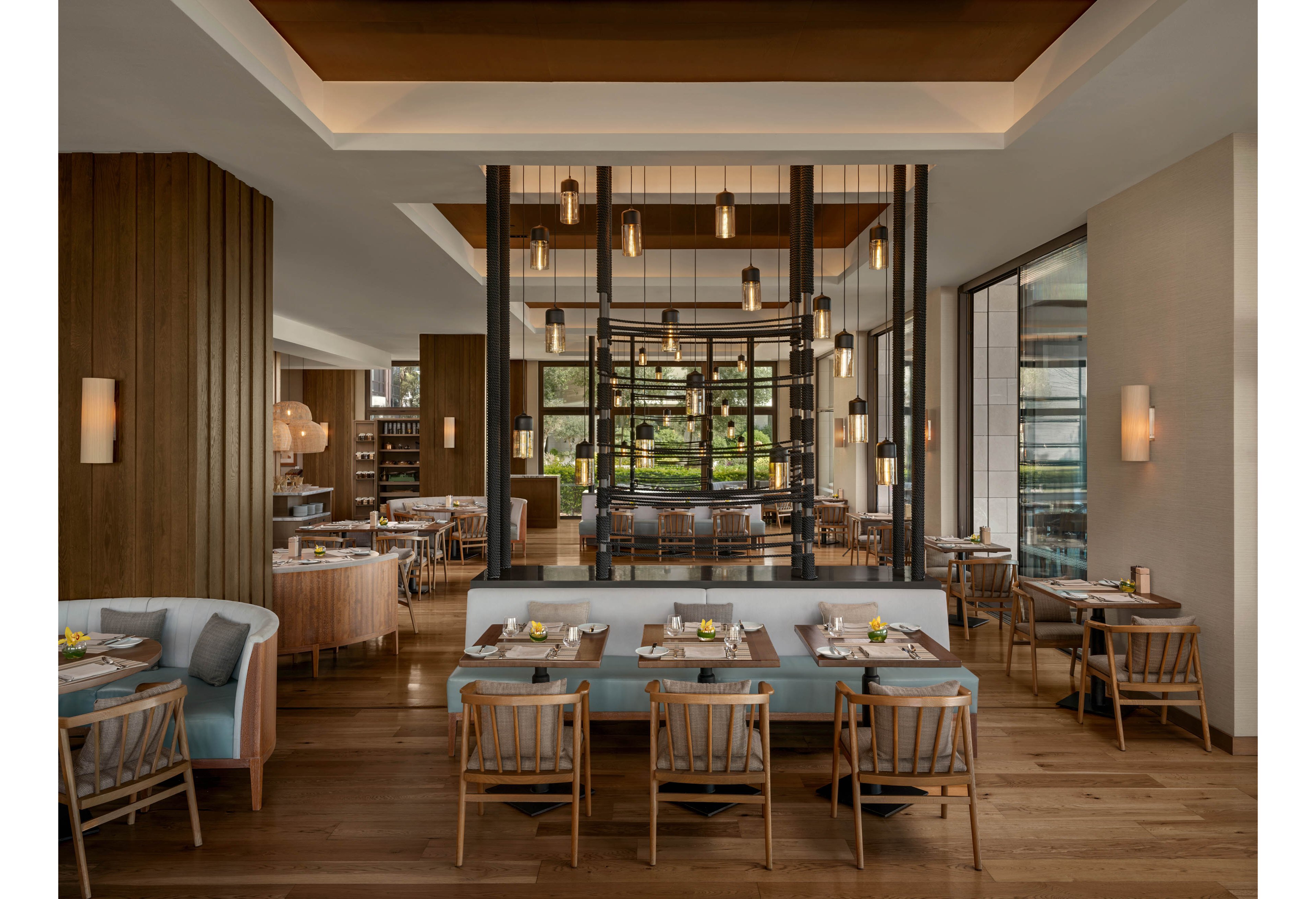 A contemporary restaurant dining room features neatly arranged tables and wooden chairs in a warm, inviting setting. Large windows allow natural light to illuminate the space, highlighting the neutral tones and soft blue upholstery. Pendant lights hang from the ceiling above a central glass partition, adding a stylish focal point. The overall atmosphere is calm, refined, and suitable for fine dining or casual gatherings.