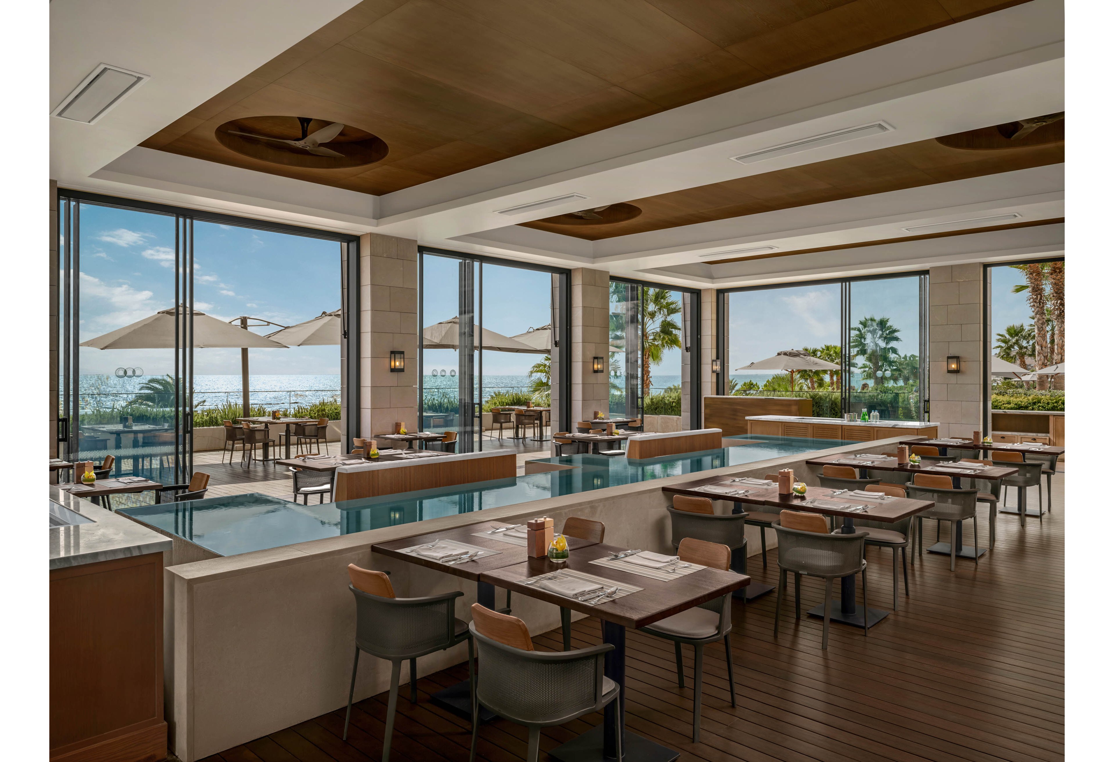 A contemporary restaurant interior features neatly arranged tables and chairs beside sleek rectangular water features. Large floor-to-ceiling windows open to a terrace with umbrellas and views of the ocean and palm trees. Warm wood tones, neutral upholstery, and clean architectural lines create a sophisticated yet relaxed dining atmosphere. The space appears bright and airy, ideal for daytime meals or casual gatherings by the sea.