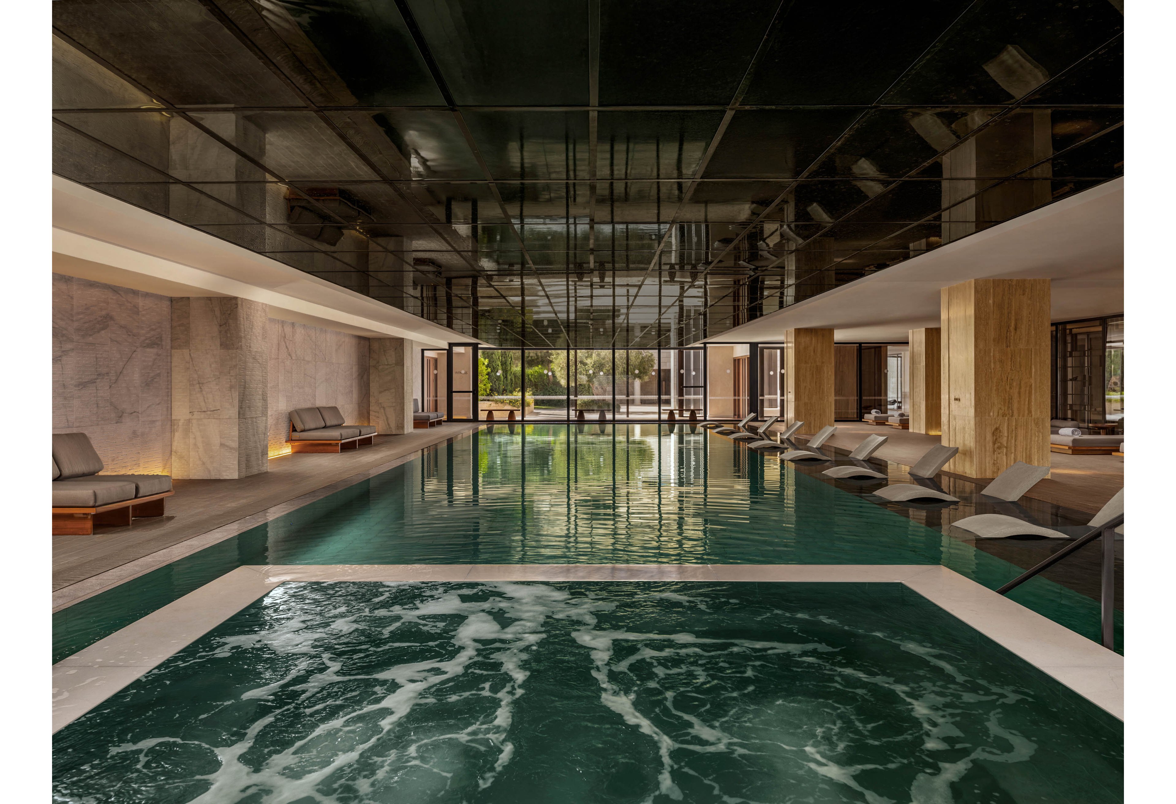A contemporary indoor swimming pool with an attached spa area is featured in this image. The space includes cushioned lounge chairs arranged neatly along both sides of the pool, creating a relaxing resort-like atmosphere. Warm wood accents, marble walls, and a reflective dark ceiling contribute to the sophisticated design. Large windows at the far end let in natural light and offer a view of greenery outside.