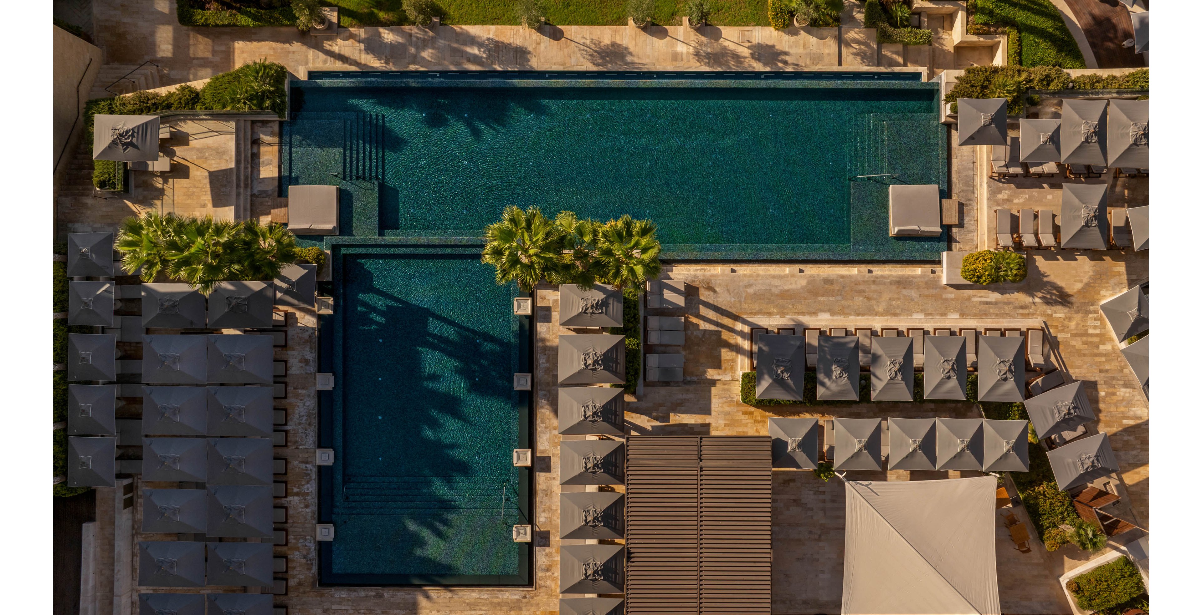 An aerial view shows two large rectangular swimming pools in a resort setting. Neatly arranged sun loungers and umbrellas line the pool decks, creating a sense of order and symmetry. Palm trees and landscaped pathways add greenery and contrast to the warm stone surfaces. The scene suggests a tranquil, upscale vacation environment under clear daylight.