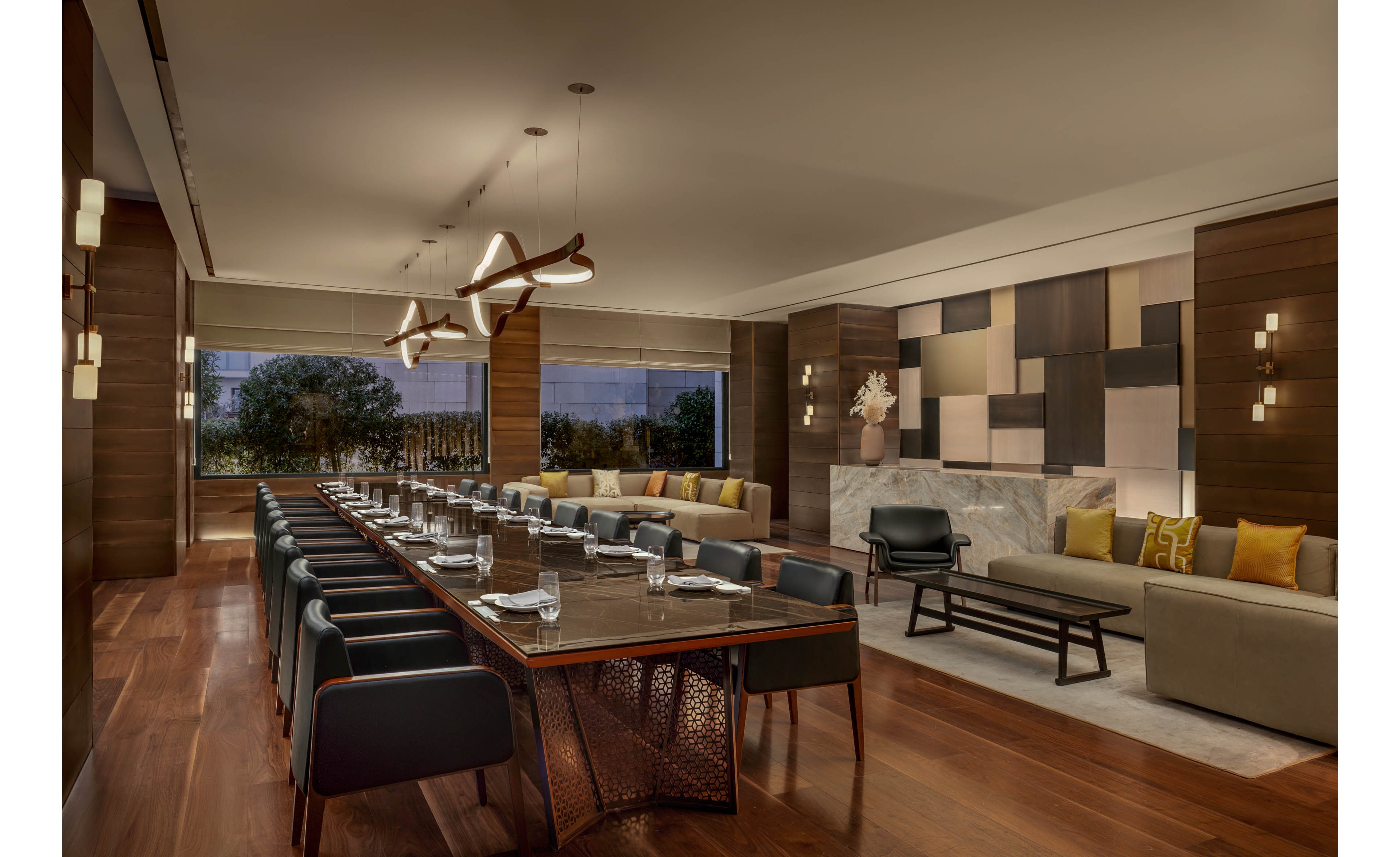 A long conference table is set for a meeting in a modern hotel-style room. The space features sleek wooden floors, contemporary pendant lighting, and upholstered chairs arranged neatly around the table. A comfortable lounge area with sofas, armchairs, and patterned wall panels sits adjacent to the meeting space. Large windows reveal greenery outside, adding a calm and sophisticated atmosphere.
