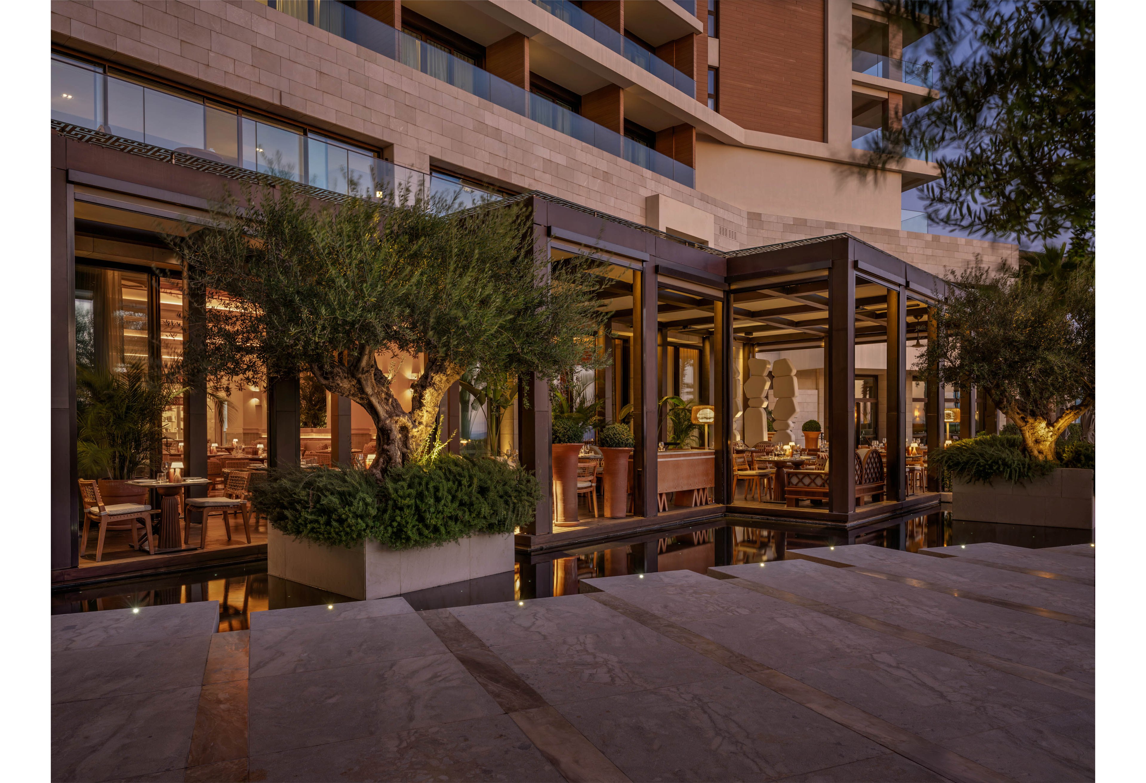 A contemporary hotel restaurant opens onto a stylish outdoor terrace at dusk. Large glass windows reveal warmly lit dining tables and wooden furnishings inside. The terrace is framed by lush potted trees and greenery, reflecting softly in a shallow water feature. The building’s modern architecture and layered balconies rise above the inviting dining area, creating an upscale yet relaxed atmosphere.