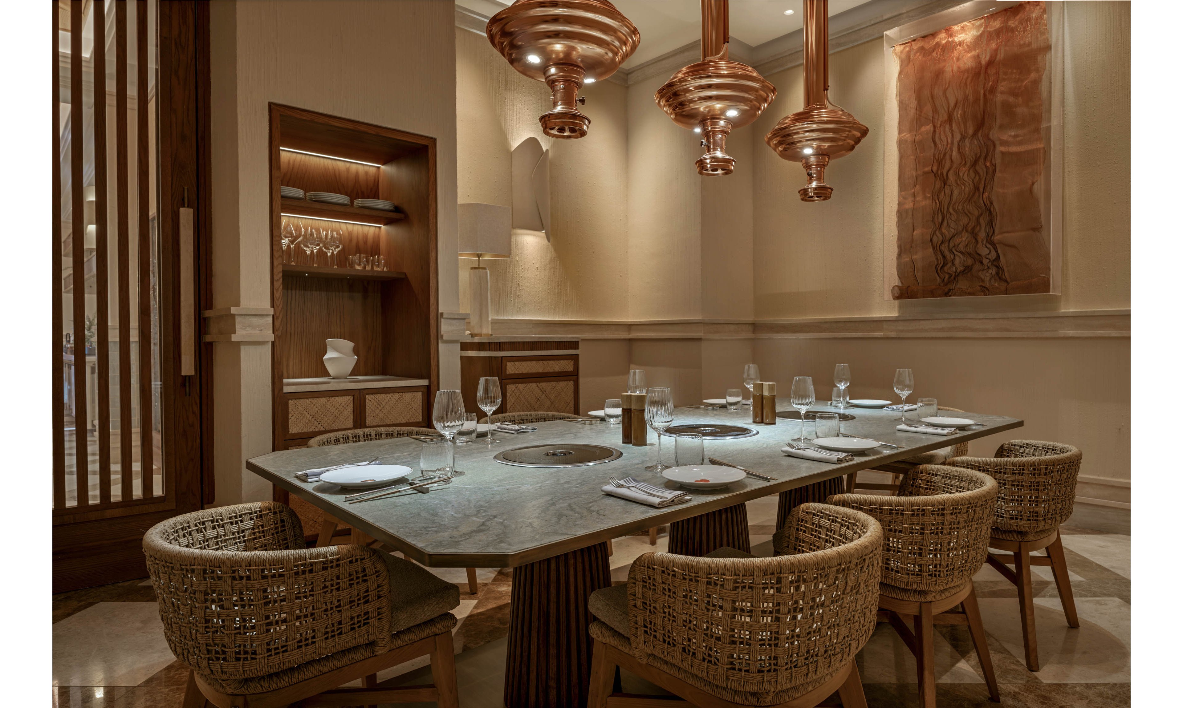 A sophisticated private dining room features a stone-topped table set for six guests. Woven armchairs surround the table, which is arranged with plates, wine glasses, and folded napkins. Warm copper pendant lights hang above, casting a soft glow over the neutral-toned interior. Built-in shelving with glassware and subtle wall art enhance the upscale, intimate atmosphere.