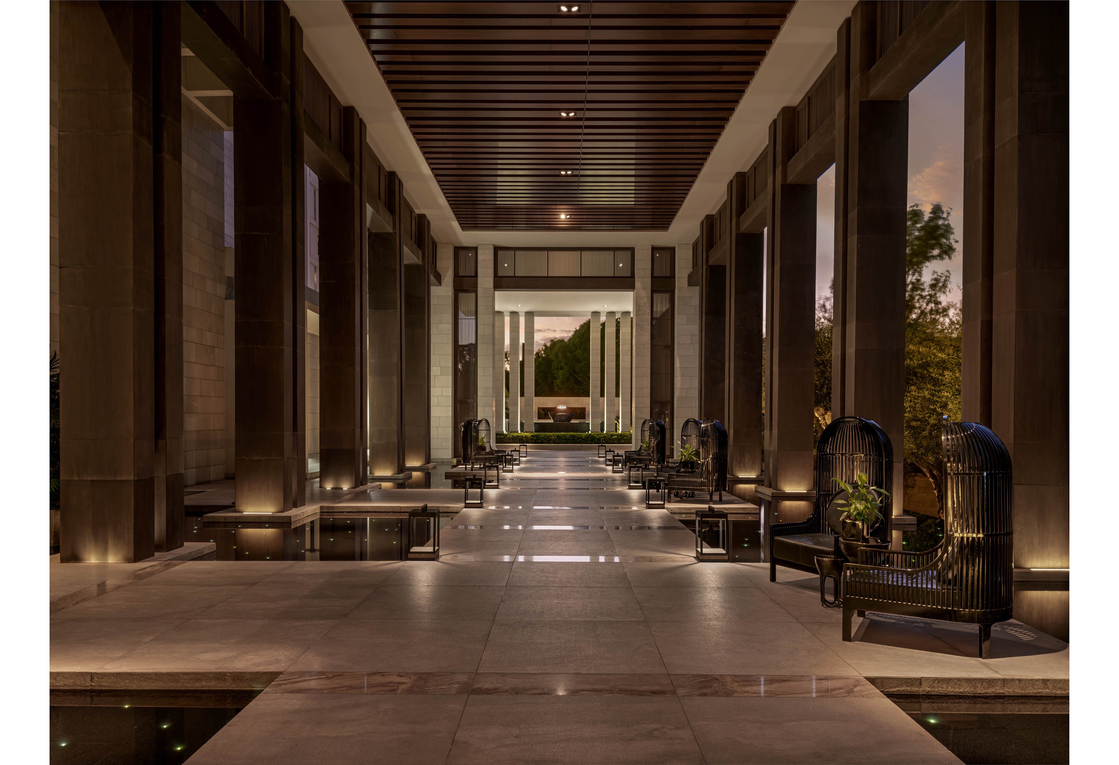 A grand open air lobby features tall columns and a long central walkway leading toward a garden view. Elegant lounge chairs and small tables line both sides of the corridor, with decorative plants adding greenery. Warm architectural lighting highlights the stone flooring, dark structural beams, and reflective water features. The scene is captured at dusk, creating a tranquil and sophisticated atmosphere.