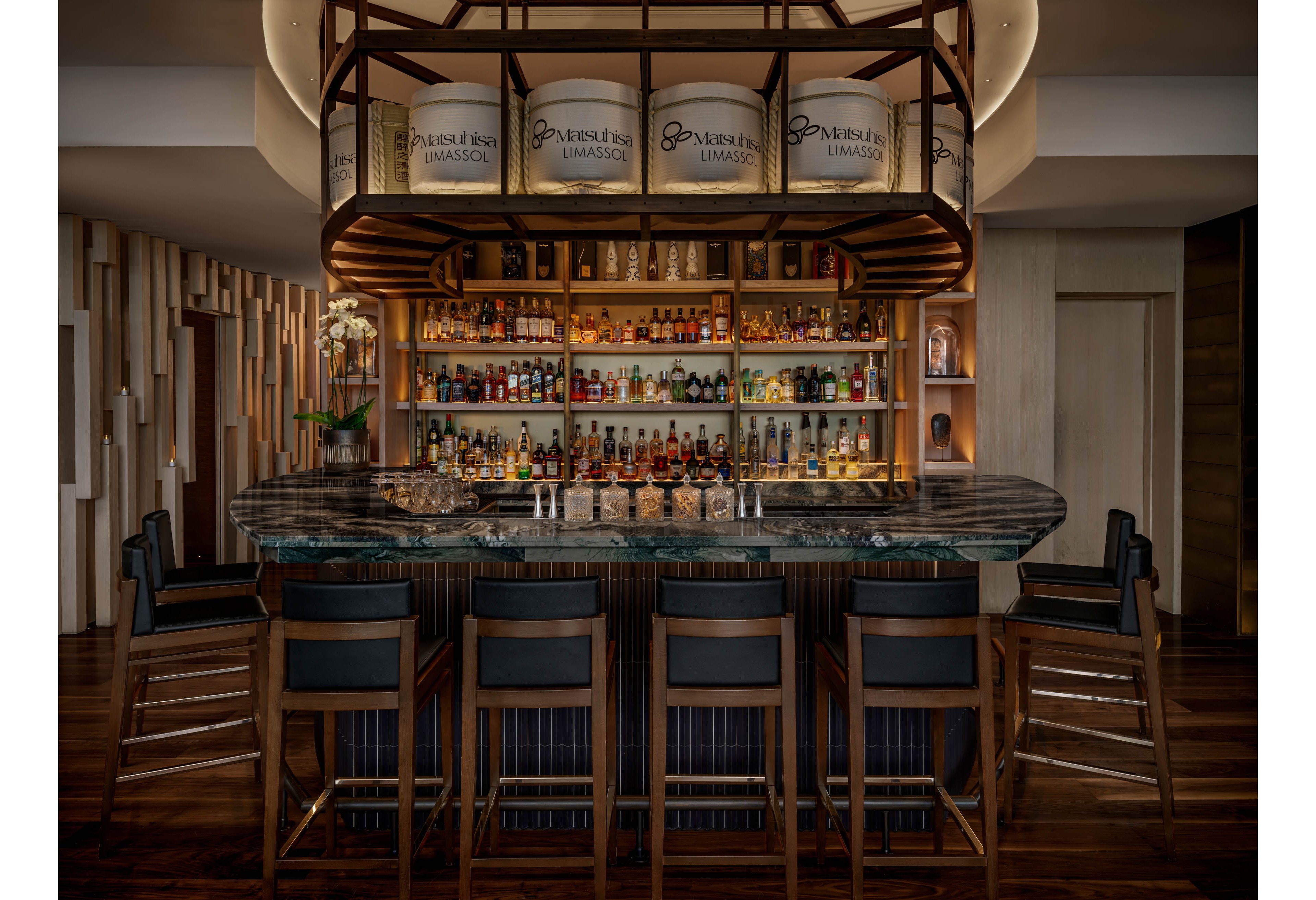 A sophisticated hotel bar features a dark marble counter surrounded by high wooden bar stools. Behind the counter, shelves are filled with an extensive selection of liquor bottles and glassware, all warmly lit. A decorative overhead structure frames the bar, with cylindrical panels displaying the words 'Limassol' and other partially visible text. The ambiance is intimate and upscale, with rich wood tones and soft lighting creating a cozy evening atmosphere.