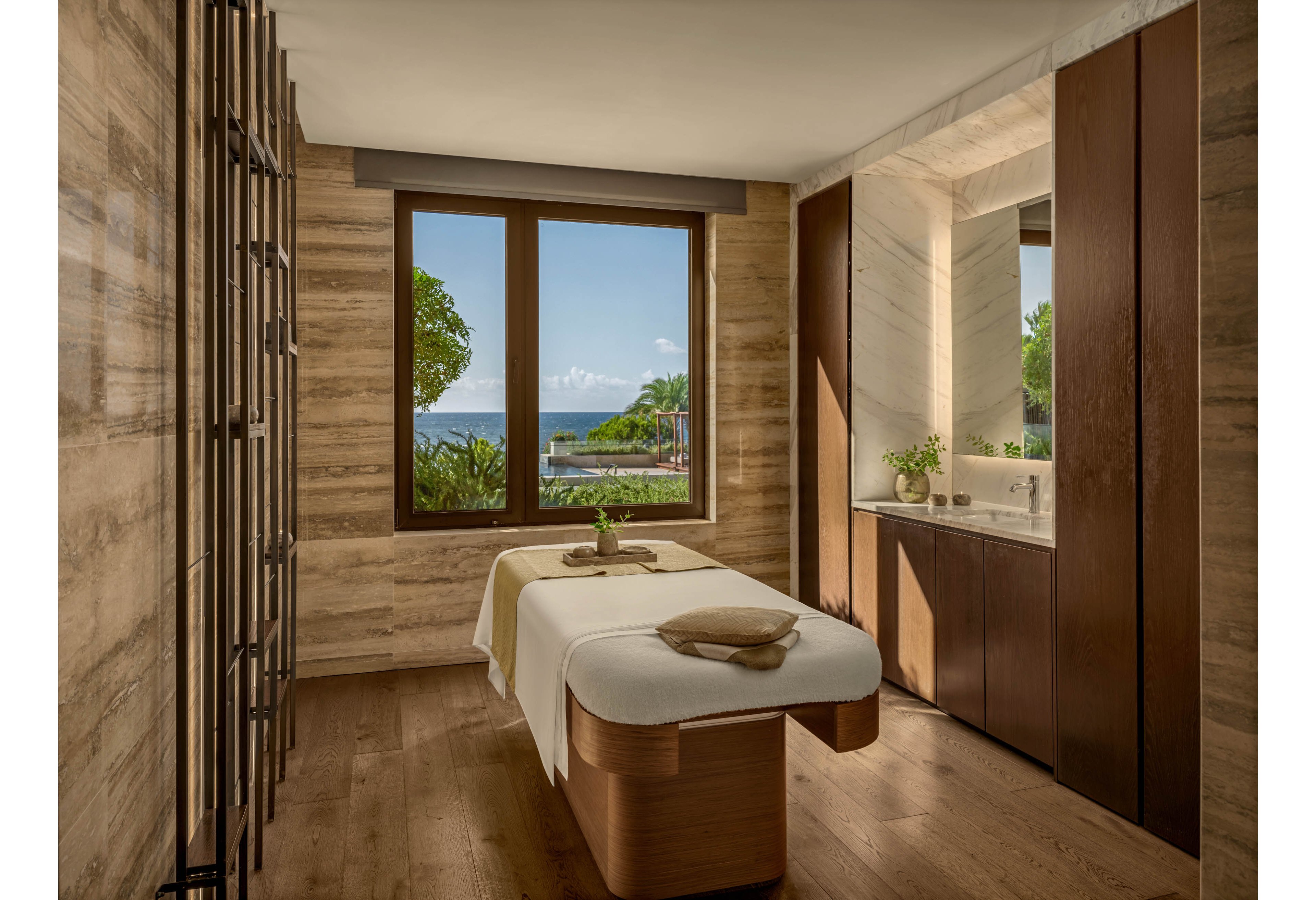 A serene spa treatment room features a massage table neatly prepared with folded towels and a small plant centerpiece. Large windows frame a lush garden and a distant ocean view under a bright sky. Warm wooden floors and beige stone walls create a natural, calming atmosphere. Built-in cabinetry and a countertop with a sink add a modern, minimalist touch to the tranquil space.