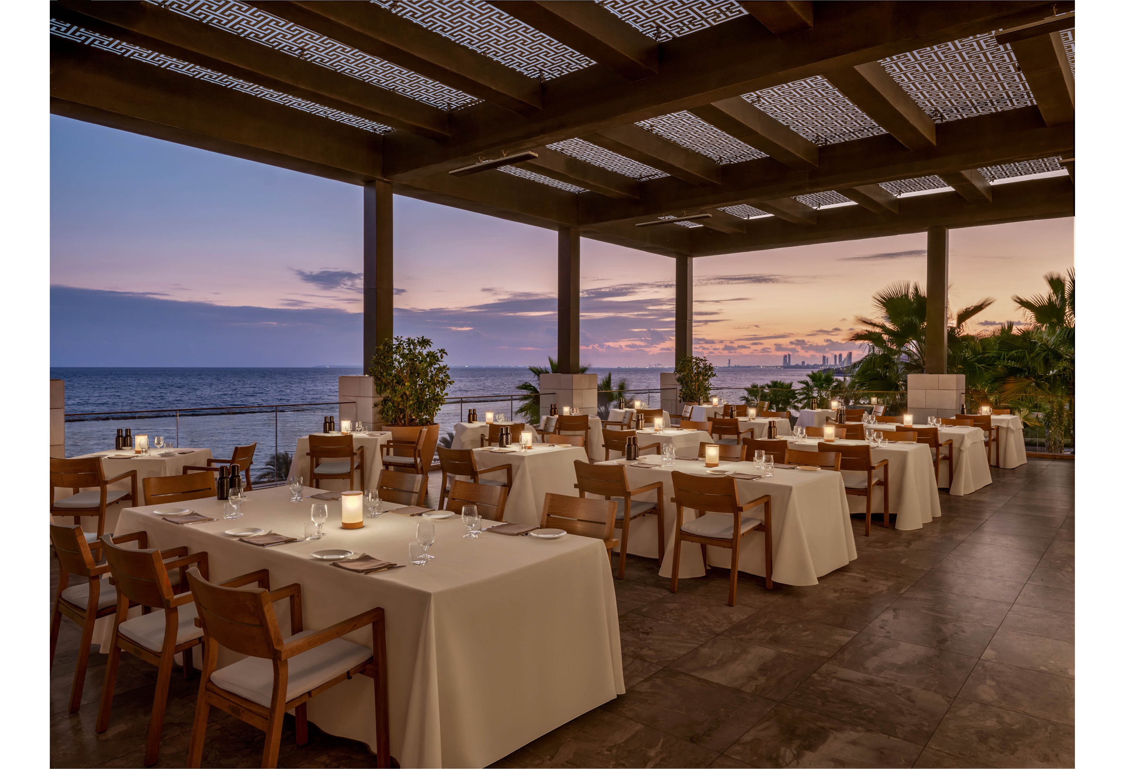An open-air restaurant terrace is set with neatly arranged tables covered in white linens. Soft candlelight glows on each table, creating a warm and inviting atmosphere. The space overlooks a calm ocean with a colorful sunset sky and distant city skyline. Modern wooden chairs, potted plants, and a partially covered ceiling add to the sophisticated coastal ambiance.
