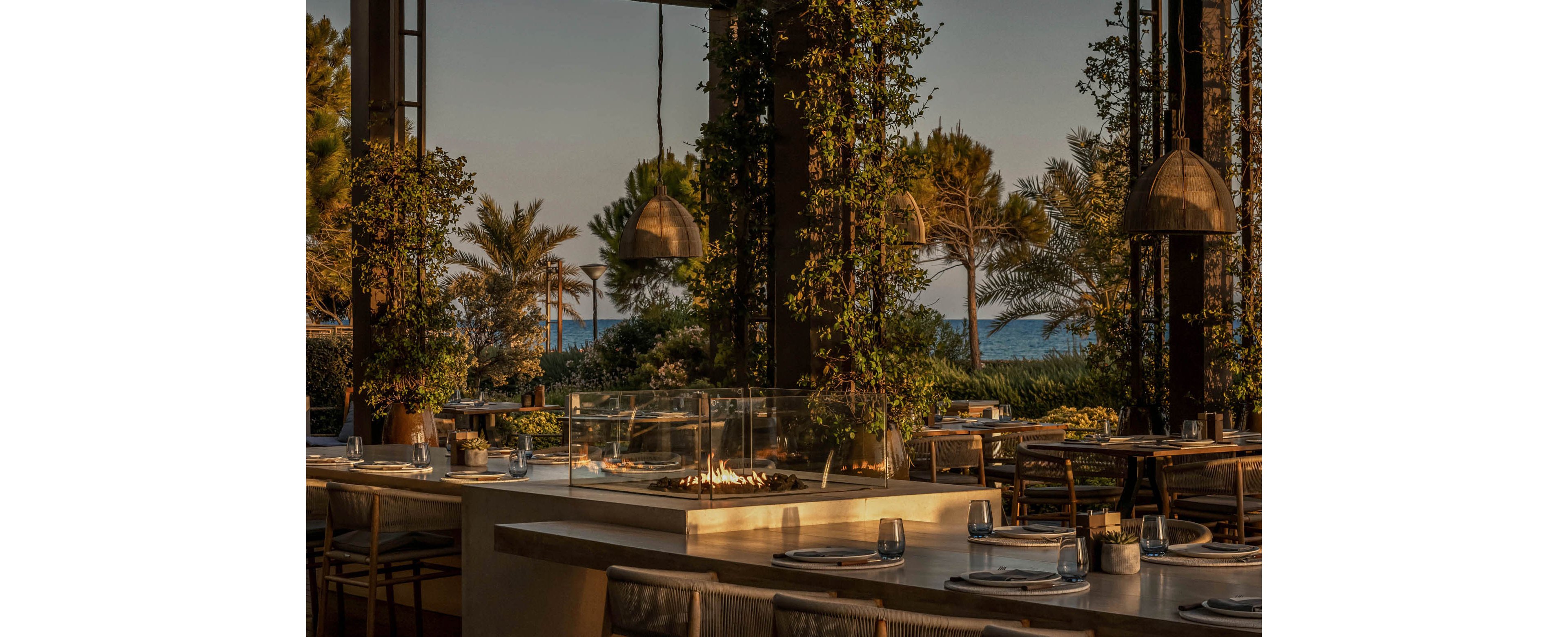 An elegant open-air restaurant terrace is shown at sunset, featuring long stone-topped tables and neatly arranged chairs. Warm pendant lights hang from the ceiling, casting a soft glow over table settings and a central linear fire feature. Lush climbing plants wrap around structural columns, framing views of palm trees and distant water. The overall atmosphere feels tranquil and sophisticated, ideal for evening dining in a resort-like setting.