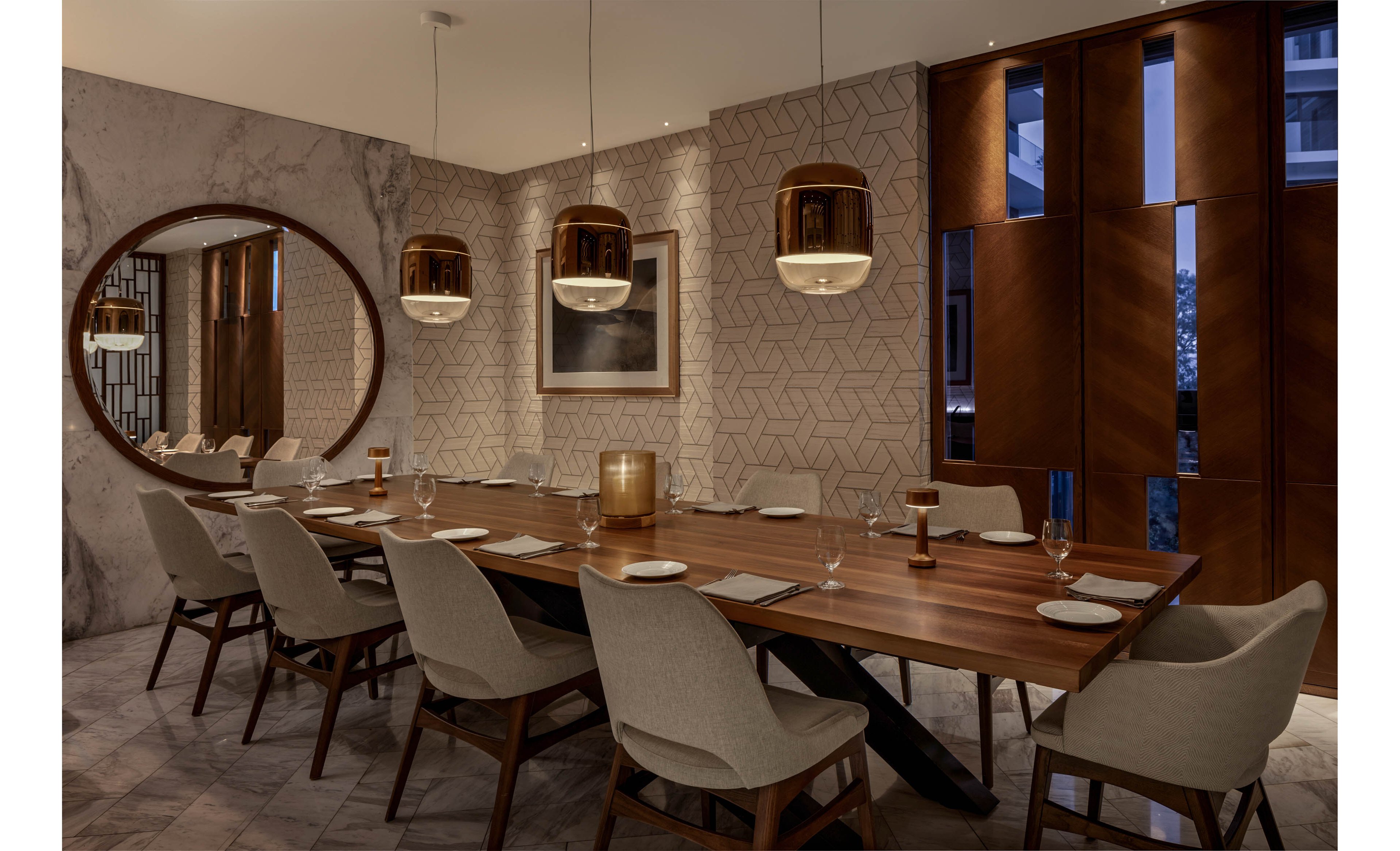 A contemporary dining room features a long wooden table set with plates, glasses, and napkins. Neutral upholstered chairs line both sides of the table, creating an inviting space for a group meal. Warm pendant lights hang above, casting a soft glow on the tabletop and highlighting the rich wood tones. Geometric patterned walls, a large round mirror, and sleek paneling contribute to the sophisticated, modern ambiance.