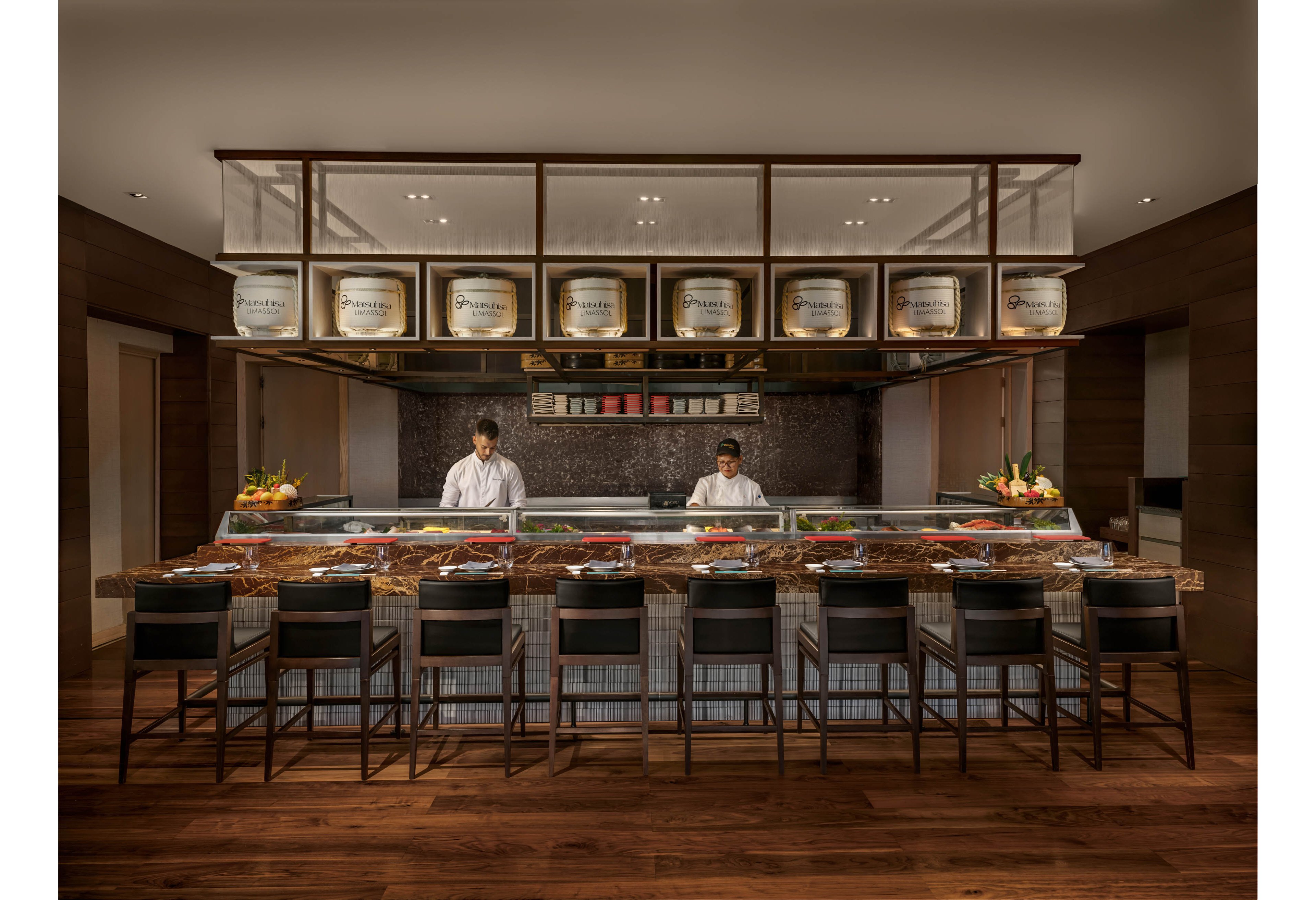 A contemporary sushi bar features a long stone counter lined with high black chairs. Two chefs stand behind the counter preparing dishes, with colorful plates of sushi and fresh ingredients displayed in front of them. Wooden flooring and warm lighting create an inviting atmosphere, while decorative barrels and shelves are arranged above the workspace. The overall setting suggests a refined, upscale dining experience in a Japanese restaurant.