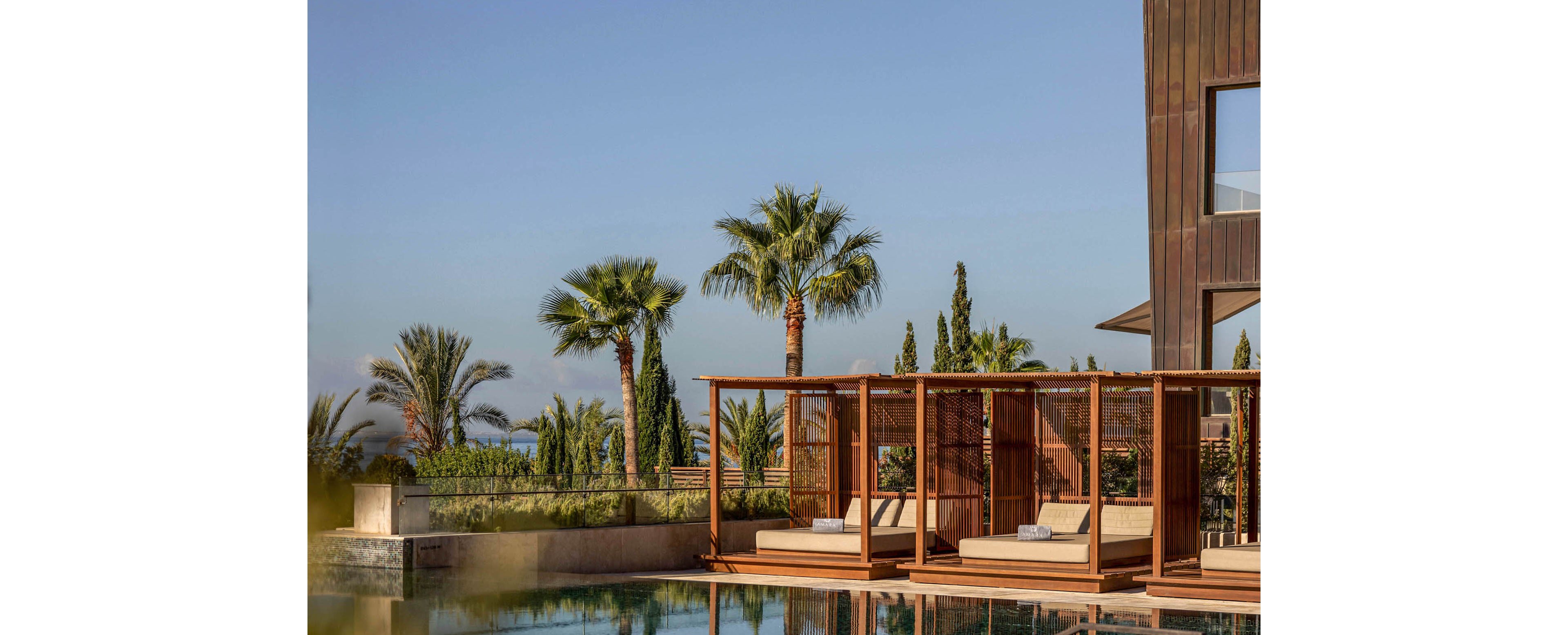 A contemporary resort-style swimming pool reflects a clear blue sky and surrounding palm trees. Wooden cabanas with lounge chairs line the poolside, creating a luxurious and relaxing atmosphere. Part of a modern building with clean architectural lines is visible on the right side of the image. The scene suggests a warm, tranquil outdoor setting ideal for vacation or leisure.