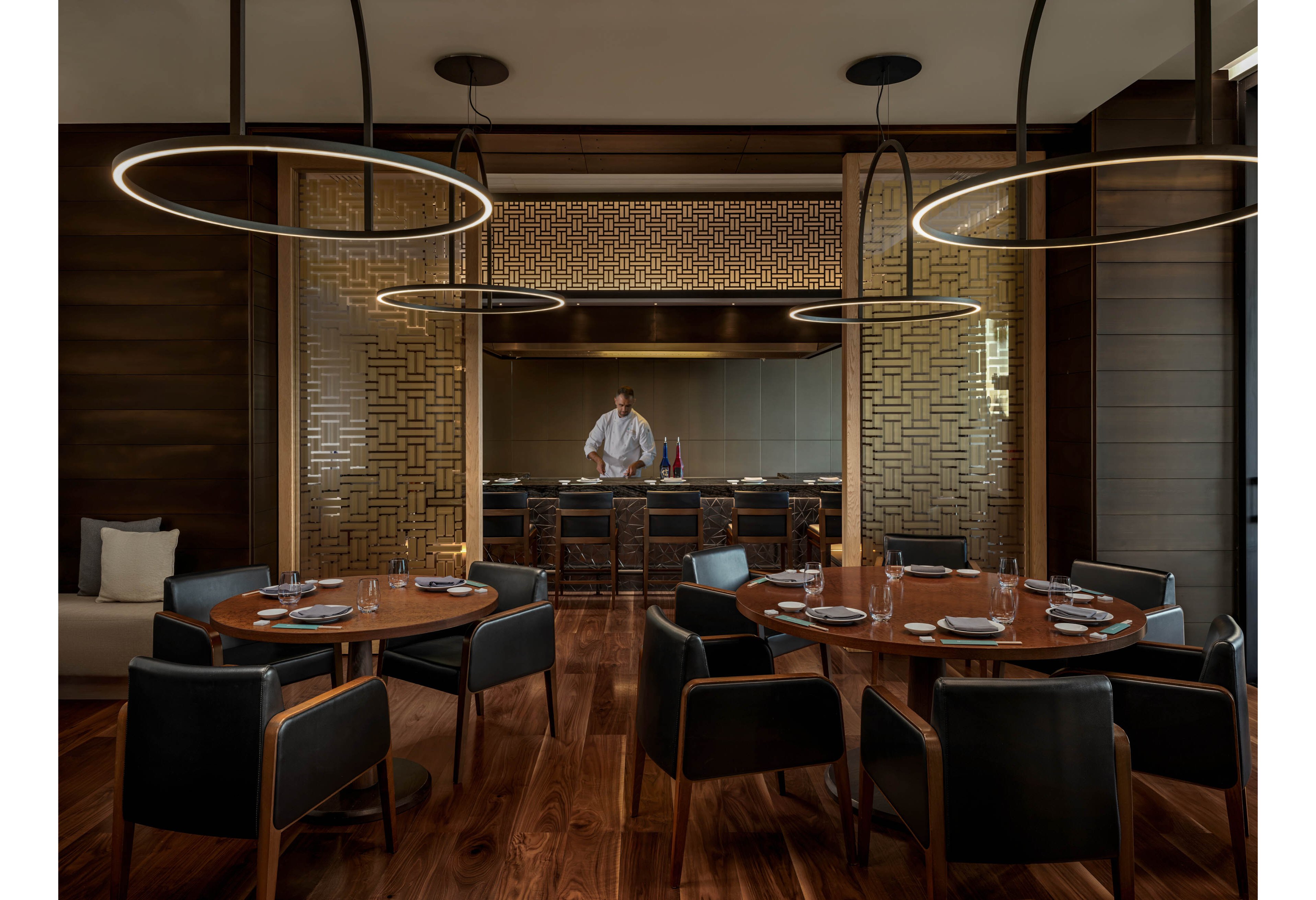 A contemporary fine dining restaurant interior features round wooden tables set with plates, glasses, and cutlery. Dark upholstered chairs surround the tables on a polished wooden floor, creating an intimate seating area. In the background, a chef stands behind a counter in an open kitchen framed by decorative golden lattice panels. Circular pendant lights hang from the ceiling, casting a warm, sophisticated glow over the space.