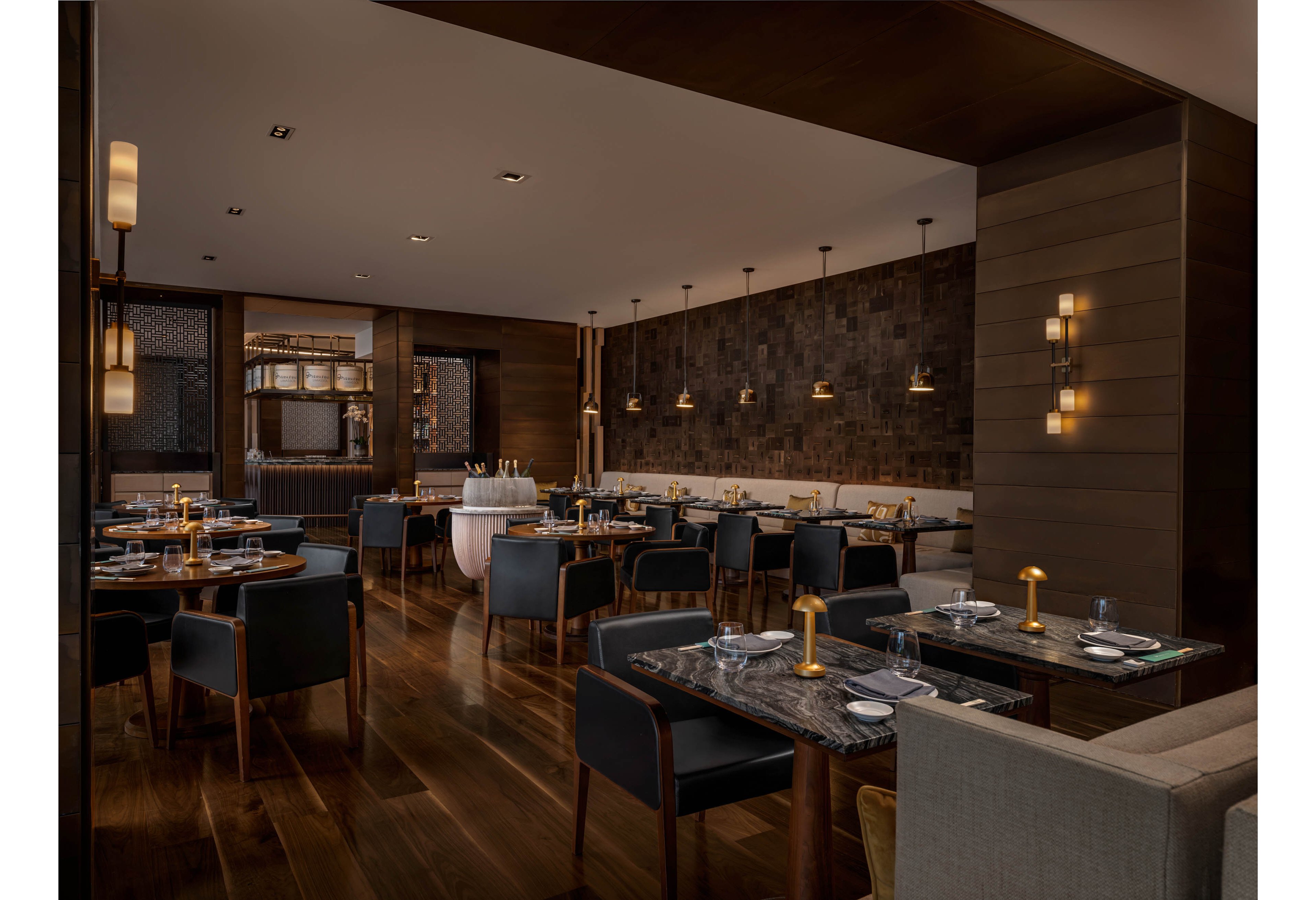 A contemporary restaurant dining room features neatly arranged tables and chairs on a polished wooden floor. Warm ambient lighting from wall sconces and pendant lamps creates an inviting atmosphere. Place settings with plates, glasses, and cutlery are prepared on dark stone tabletops, while a bar area is visible in the background. The space appears quiet and empty, emphasizing its modern design and cozy mood.