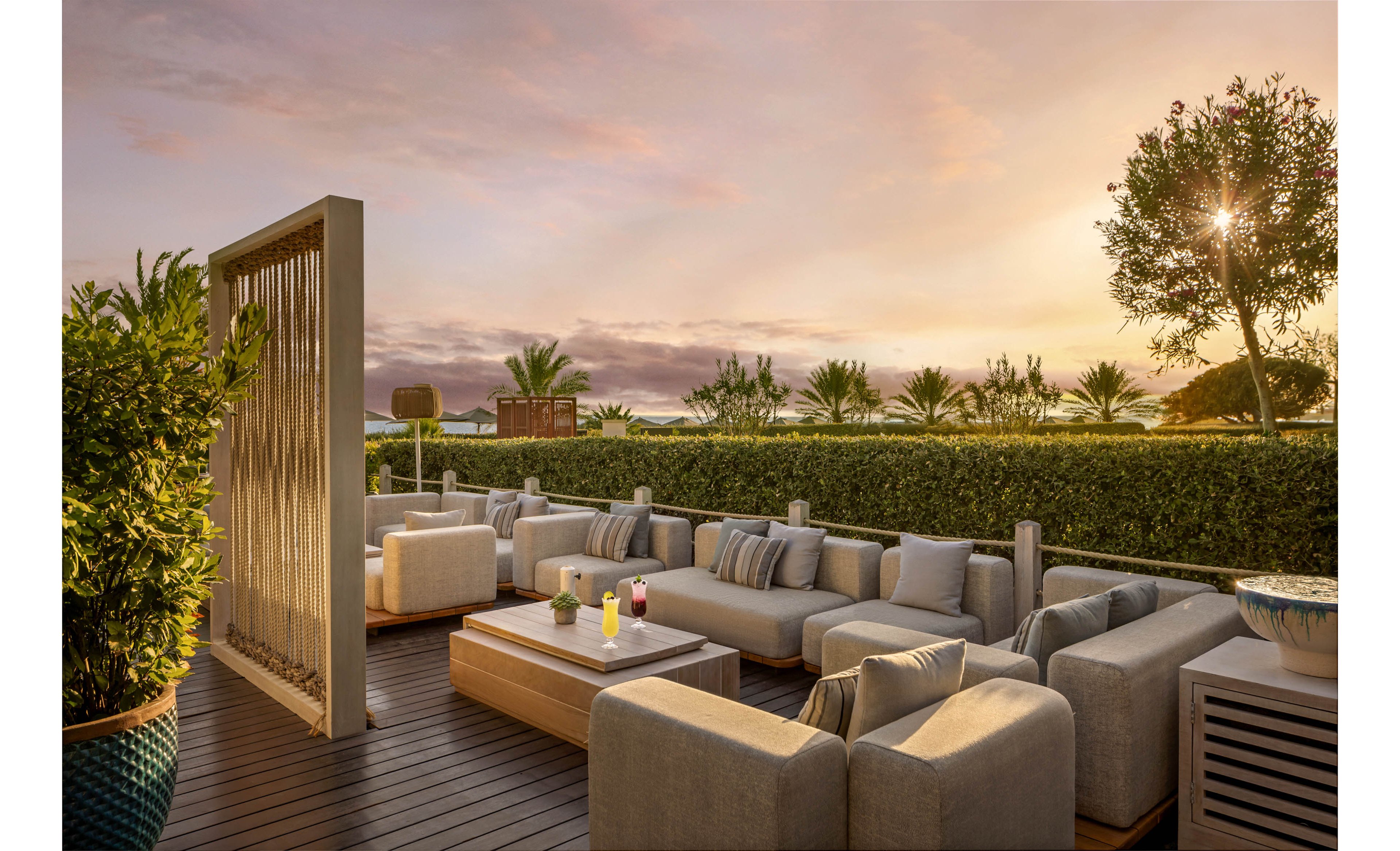 A modern outdoor lounge area features plush beige sofas arranged around a low coffee table on a wooden deck. Colorful drinks sit on the table, adding a bright accent to the neutral furniture. Lush greenery, hedges, and palm trees surround the terrace, creating a private and tranquil atmosphere. The warm sunset sky casts a golden light over the scene, emphasizing the relaxed, resort-like setting.