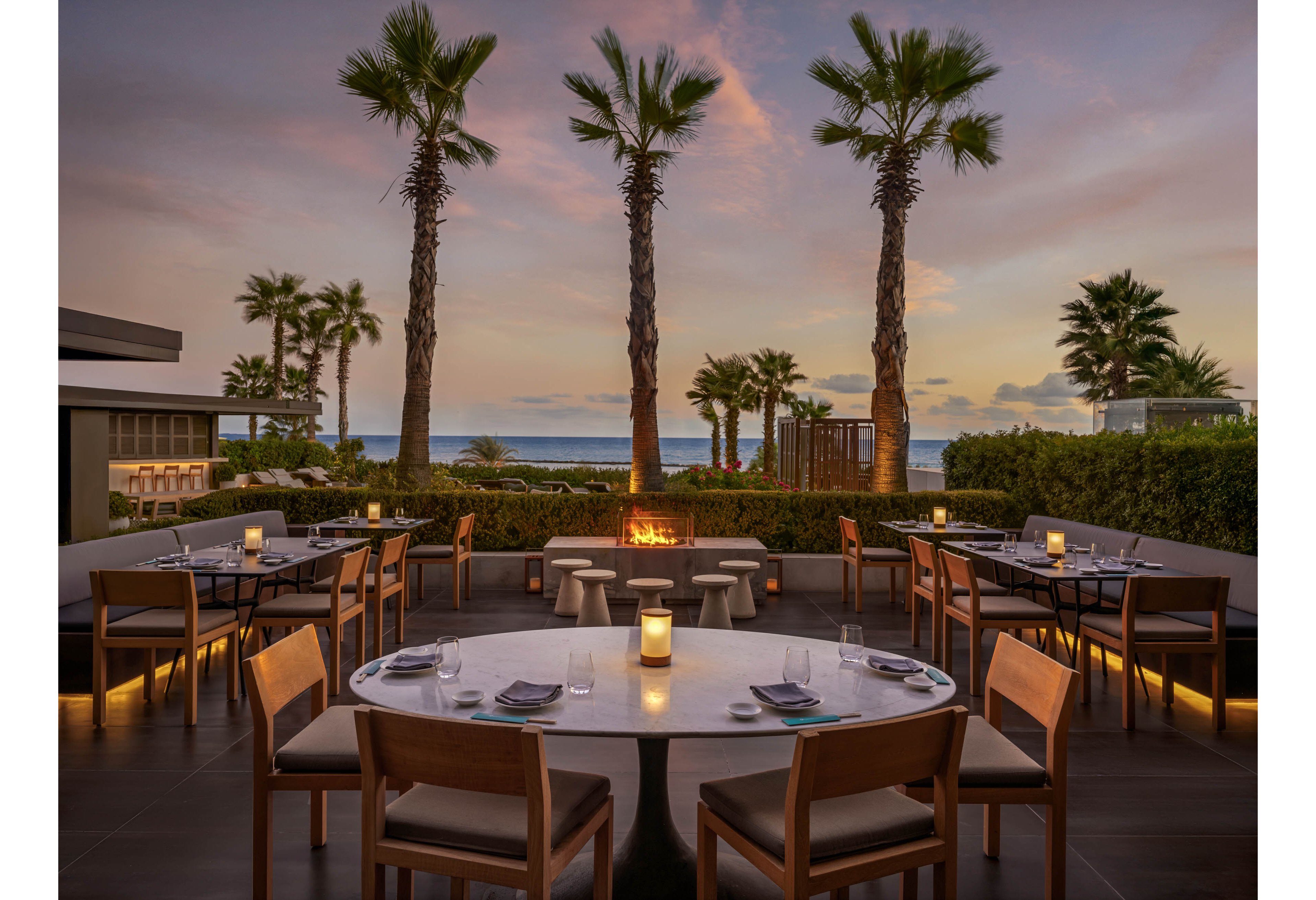 An elegant open-air restaurant terrace overlooks the ocean at sunset. Round and rectangular tables are neatly set with plates, napkins, and glassware, while a central firepit glows warmly. Palm trees frame the view of the sea and pastel sky, creating a relaxed coastal ambiance. Soft lighting from table candles and built-in fixtures enhances the sophisticated yet inviting atmosphere.