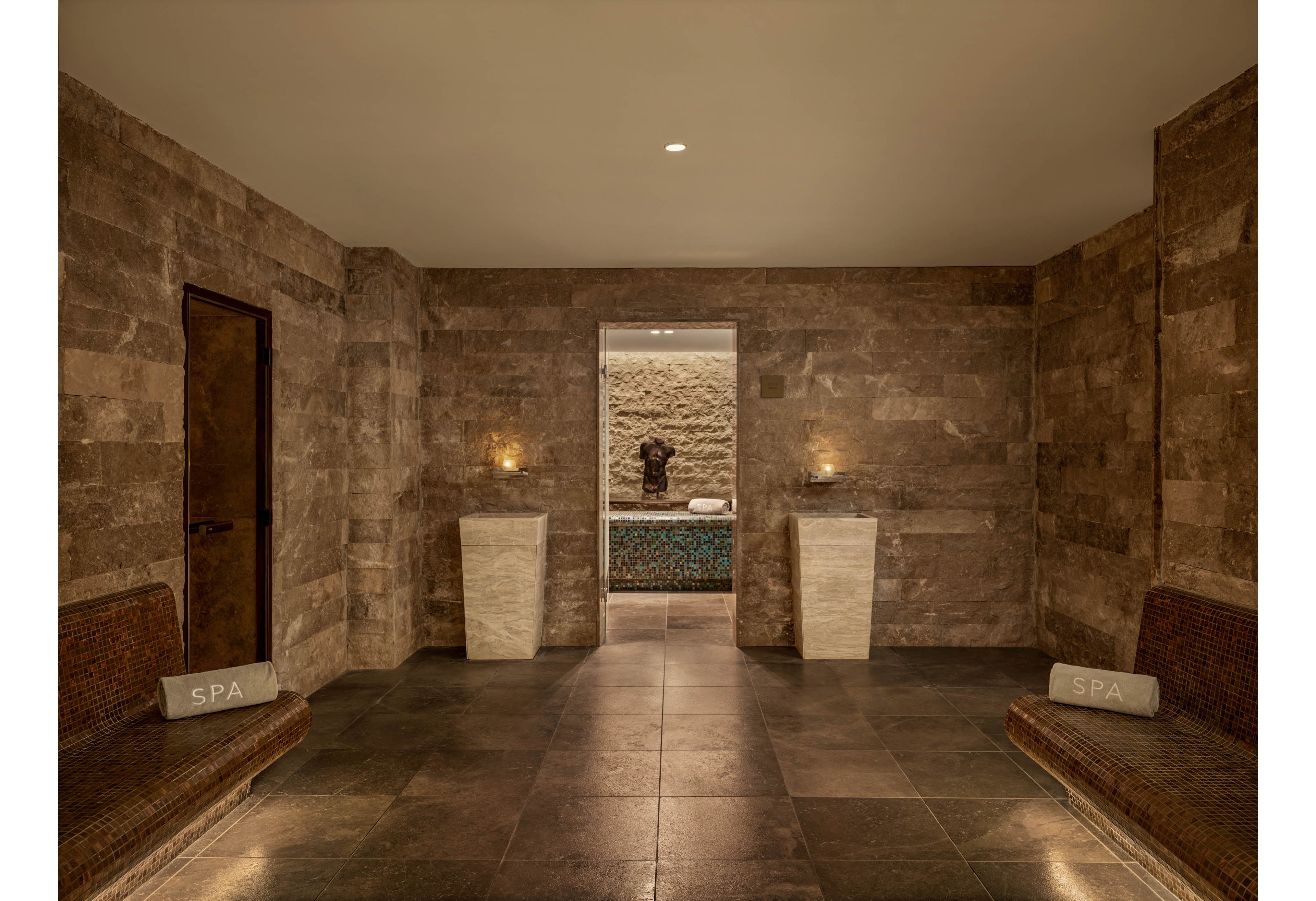 A serene spa corridor features warm stone walls and floors leading to a softly lit treatment area. Two cushioned benches line the sides, each with a pillow labeled SPA. Minimalist pedestal tables with candles enhance the calm ambiance. The neutral color palette and subtle lighting create a relaxing, luxurious atmosphere.
