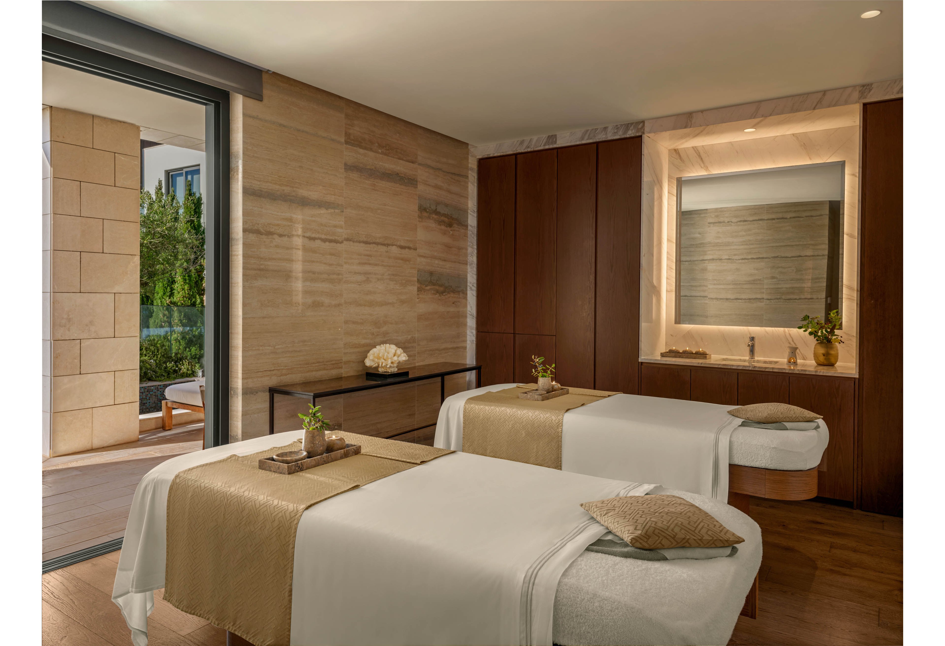 A serene spa treatment room features two massage tables neatly arranged side by side. Neutral beige and brown tones create a calming atmosphere, complemented by soft lighting and minimalist decor. A large window opens to an outdoor terrace with greenery, bringing natural light into the space. Small trays with plants and rolled towels rest on the tables, enhancing the sense of relaxation and wellness.