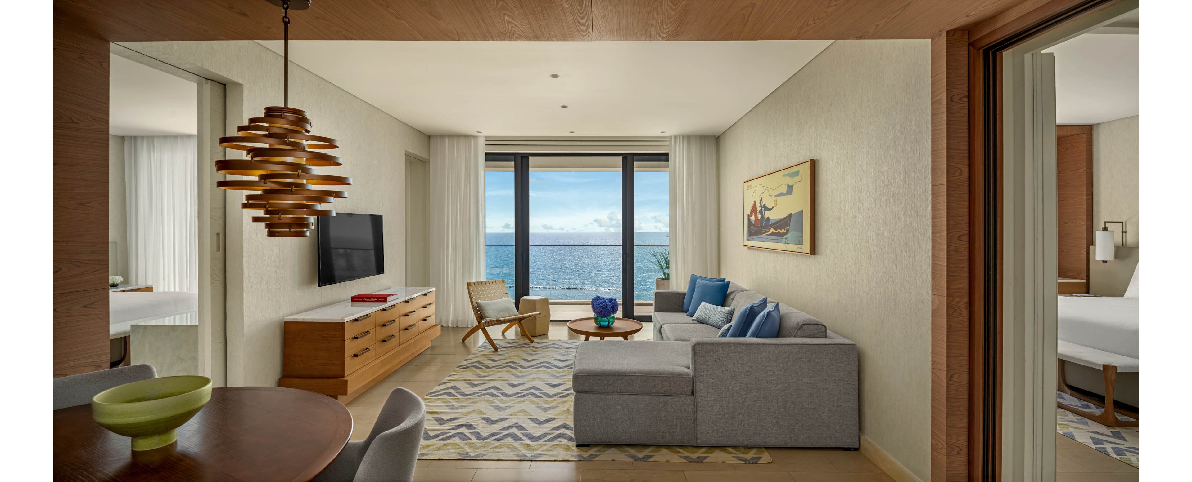 A contemporary hotel suite living room opens onto a balcony with a clear view of the ocean. The space features a gray sectional sofa with blue accent pillows, a wooden media console with a flat-screen TV, and a patterned rug. Large floor-to-ceiling windows let in abundant natural light, highlighting neutral walls and minimalist decor. Adjacent rooms with beds are partially visible, suggesting a spacious multi-room accommodation ideal for relaxation.