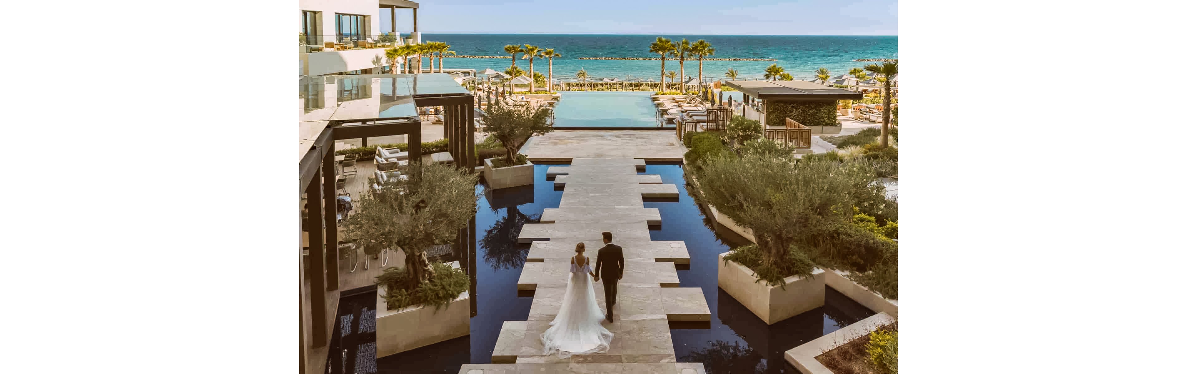 A bride and groom walk hand in hand along a modern stone pathway surrounded by reflective water features. The scene overlooks a luxurious resort pool area that opens out to a calm, turquoise ocean. Palm trees, manicured greenery, and contemporary architecture frame the romantic setting. The warm, golden lighting and clear blue sky create an elegant, dreamy atmosphere ideal for a destination wedding.