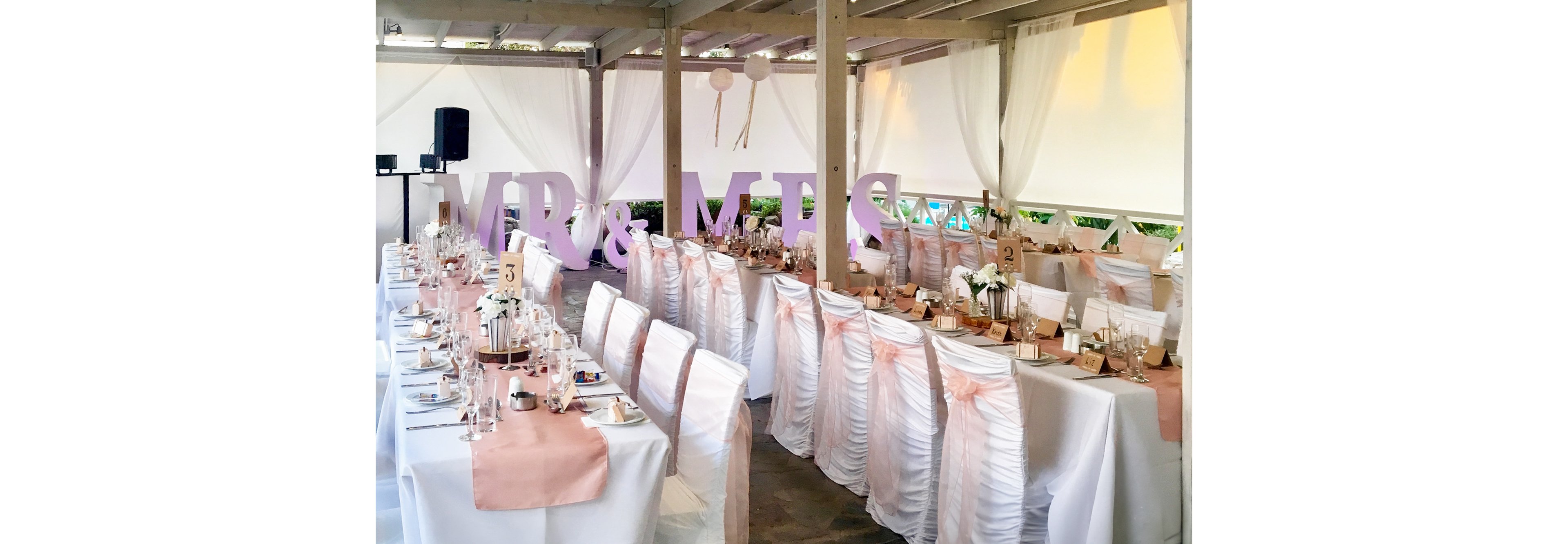 A beautifully decorated wedding reception features long banquet tables draped in white linens with soft blush pink runners. White chairs are covered with ruched fabric and tied with pink sashes, creating a cohesive and romantic look. Large freestanding letters spelling “MR & MRS” are displayed prominently near the head of the room. Table numbers, glassware, and delicate centerpieces add detail to the sophisticated indoor celebration.