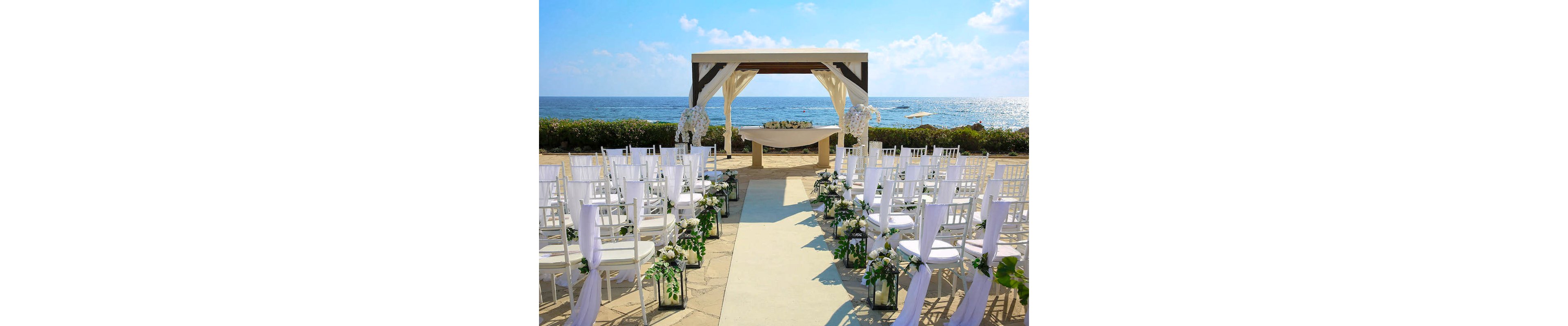 An outdoor wedding ceremony is arranged on a stone terrace overlooking the ocean. White chairs line both sides of an aisle leading to a draped gazebo-style altar. Floral arrangements and greenery decorate the aisle and the front of the structure, creating a romantic atmosphere. Bright daylight and blue sky enhance the fresh, coastal setting.