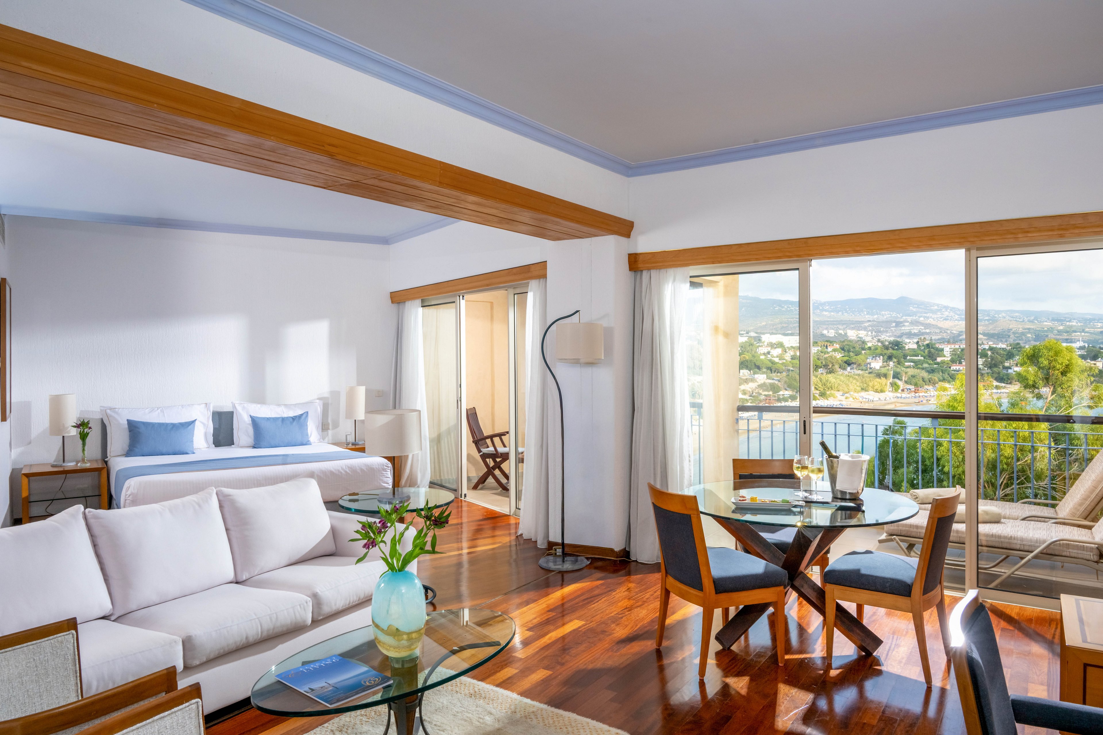 Spacious modern hotel suite features a white sofa seating area and an open-plan bedroom with a large bed. Floor-to-ceiling windows and sliding glass doors lead to a balcony overlooking water and a distant cityscape. Wooden floors, light wood furniture, and soft blue accents create a clean, coastal feel. Natural daylight fills the room, highlighting the contemporary decor and inviting atmosphere.