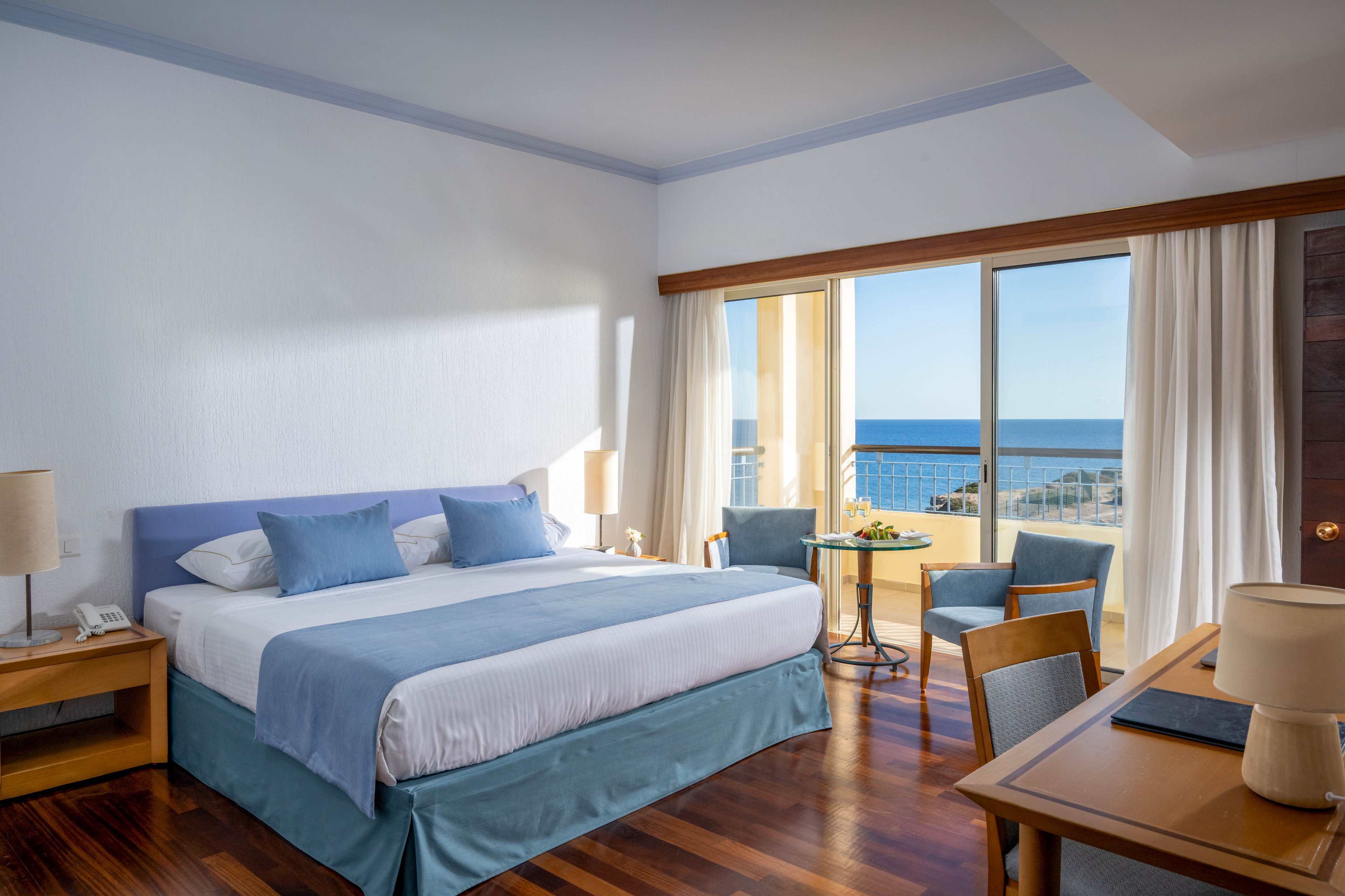 A spacious hotel bedroom features a large neatly made bed with blue and white linens. The room includes wooden furniture, such as bedside tables, a small round table with two chairs, and a work desk with a lamp. Large sliding glass doors open to a balcony overlooking the ocean and coastal buildings. Natural light fills the space, creating a calm and inviting atmosphere with soft blue accents.