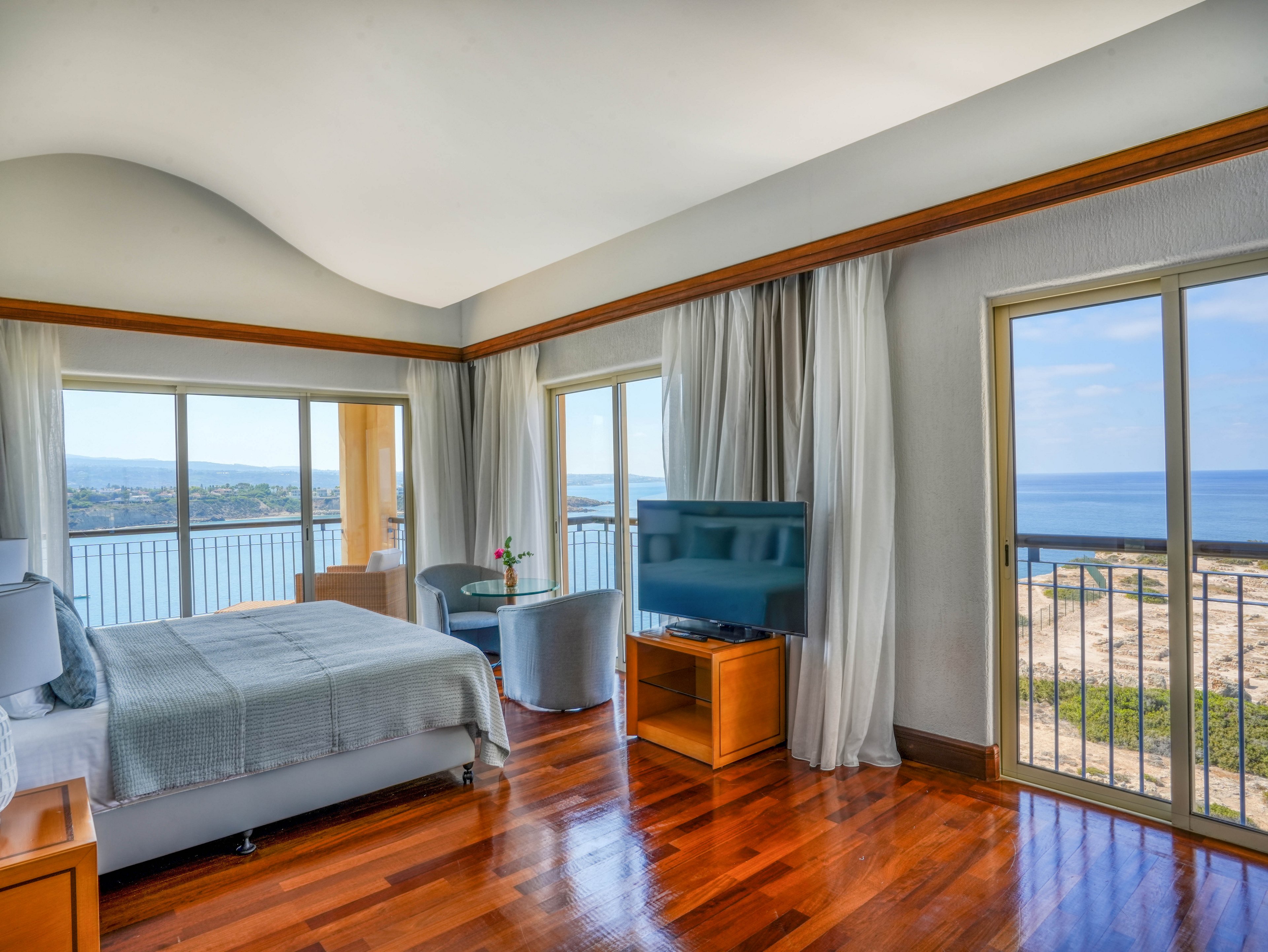 Spacious hotel bedroom featuring a large bed and polished wooden floor overlooks a scenic coastline. Floor-to-ceiling windows and sliding glass doors open to a balcony with expansive sea views. A flat-screen television sits on a wooden stand beside a small seating area with two chairs and a table holding a single flower. Soft neutral tones and abundant natural light create a calm, relaxing atmosphere.