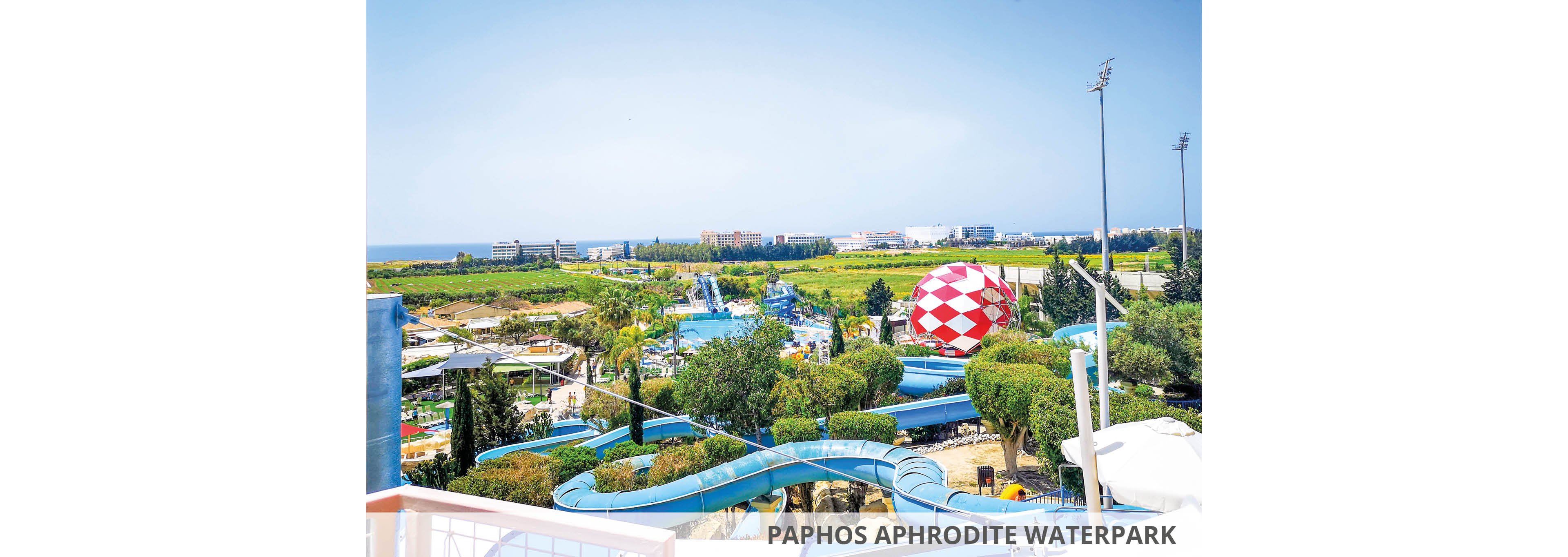 A vibrant view of Paphos Aphrodite Waterpark featuring winding blue water slides surrounded by lush greenery. The setting includes a large red and white patterned dome and distant buildings near the coastline. The image captures a sunny day with clear skies, showcasing the park's expansive layout and recreational attractions.