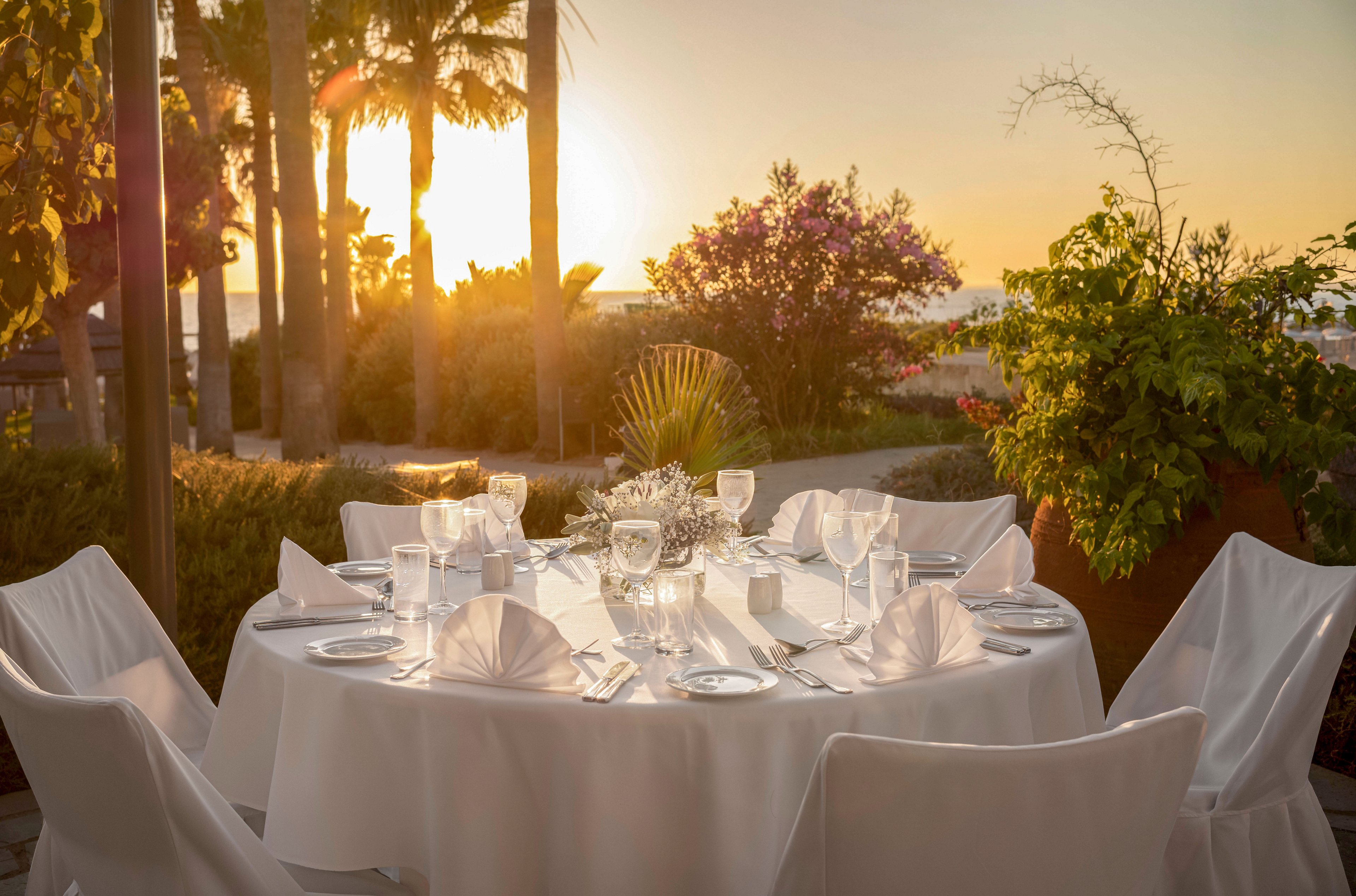 A round table is beautifully set for an outdoor dinner, covered with a crisp white tablecloth and neatly folded napkins. Glassware and plates are arranged around a floral centerpiece, suggesting a formal or celebratory occasion. The setting overlooks a garden with lush greenery, palm trees, and blooming flowers. Warm golden sunlight from the sunset creates a romantic and tranquil atmosphere.