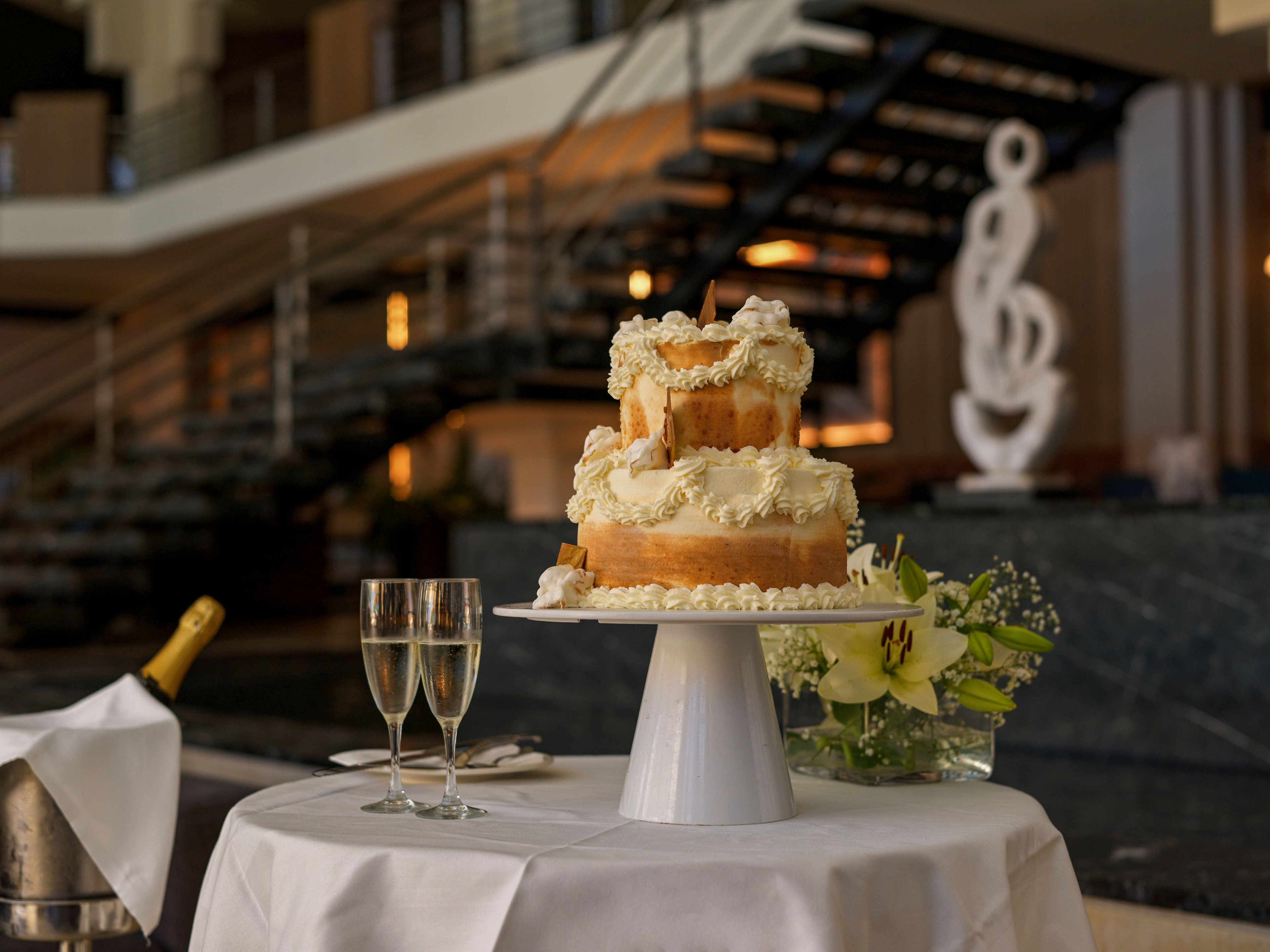 A tiered cream-covered cake is displayed on a white pedestal stand atop a round table draped in a white tablecloth. Two filled champagne glasses and a floral arrangement with white lilies sit beside the cake, suggesting a celebratory occasion. In the background, a modern staircase and abstract sculpture create a sophisticated indoor setting. Soft, warm lighting enhances the luxurious and festive atmosphere.
