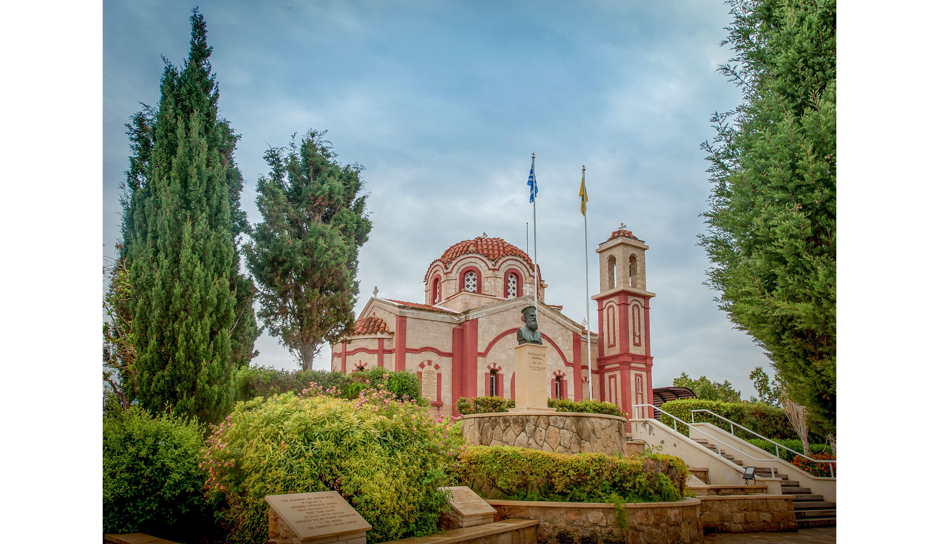 A traditional Orthodox church with red and cream detailing stands amid lush greenery and tall cypress trees. The building features a domed roof, arched windows, and an adjacent bell tower rising on the right side. In front of the church is a stone terrace with a prominent bust monument and steps leading up to the entrance. Two flags fly on poles above the monument, set against a cloudy blue sky. The overall scene feels peaceful and historic, highlighting religious architecture in a landscaped setting.